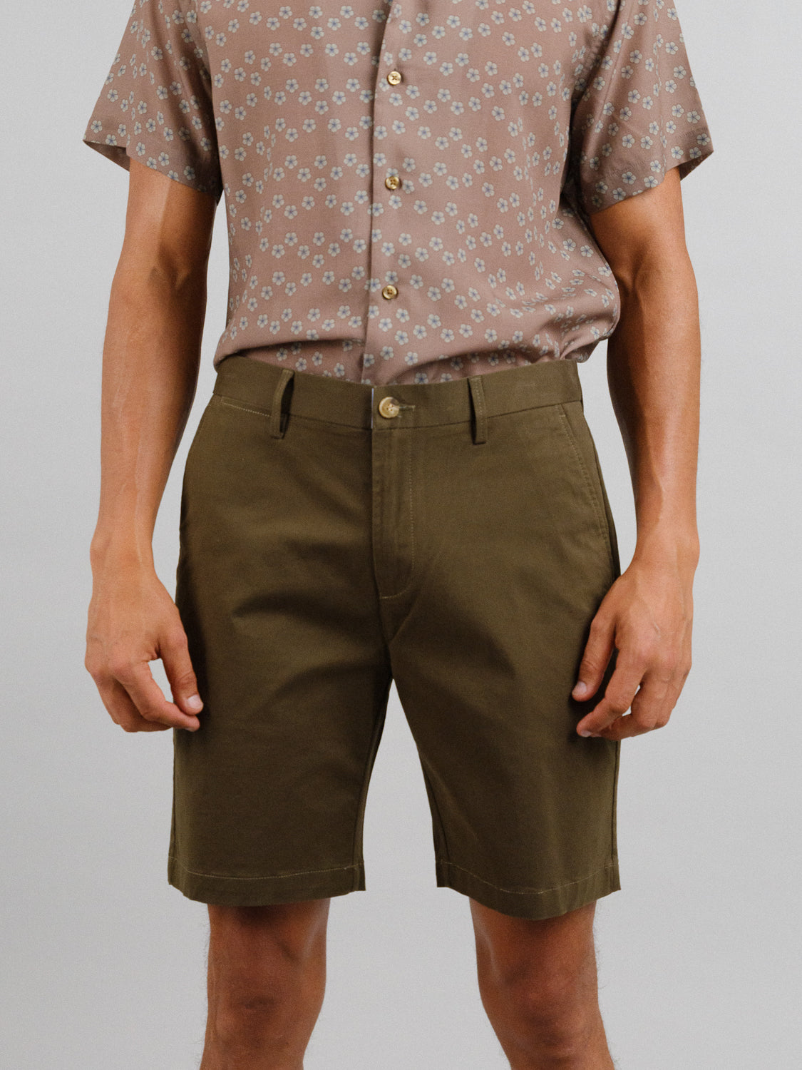 A person stands against a plain background wearing ID Menswears At Ease Relaxed Chino Short in versatile olive-green paired with a brown, short-sleeved button-up shirt featuring a small floral pattern and relaxed fit.