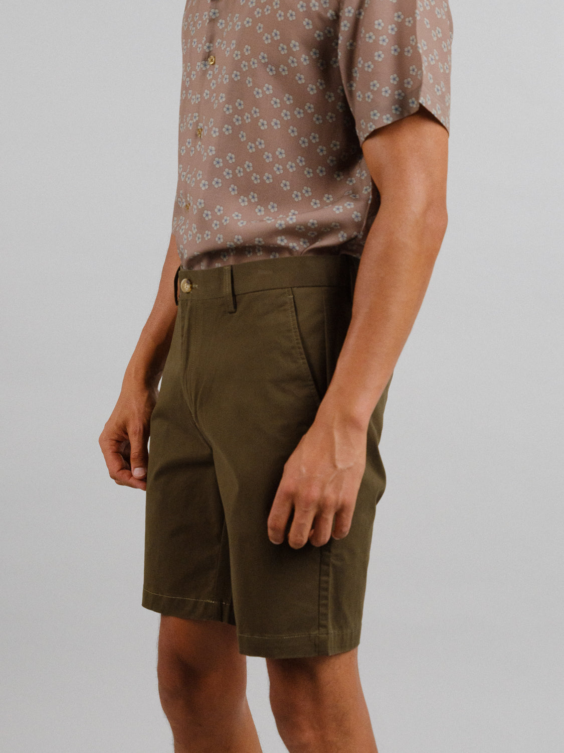 A person is shown from the shoulders down wearing a floral short-sleeve shirt and ID Menswears At Ease Relaxed Chino Short in olive green, set against a plain gray background.