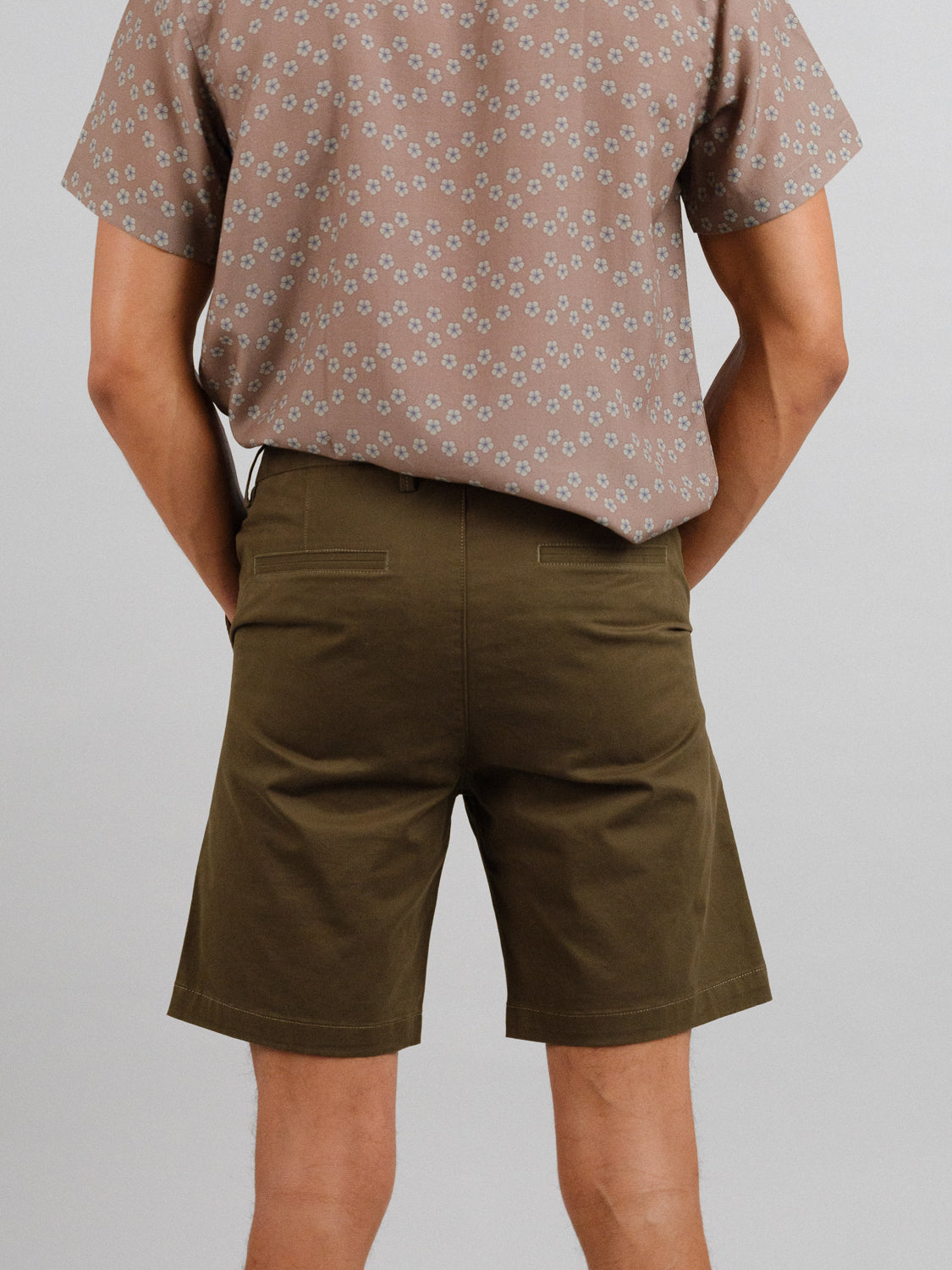 In a light gray background, a person is seen from shoulders to knees wearing ID Menswears At Ease Relaxed Chino Short in olive green and a brown patterned shirt, with their back to the camera.