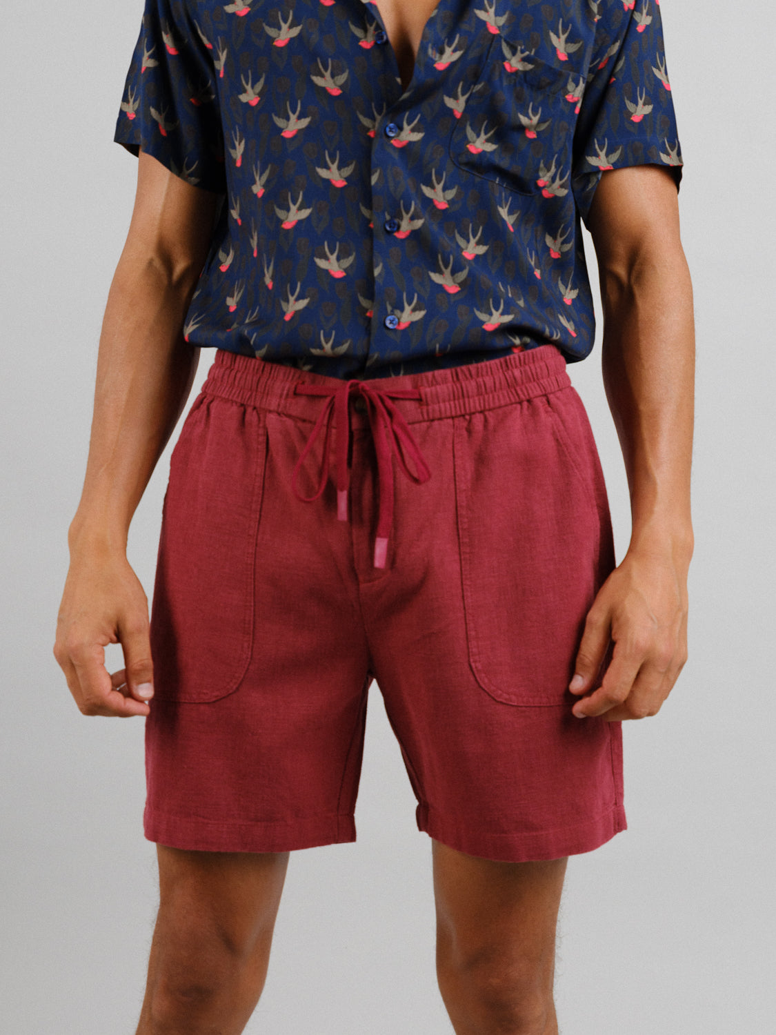 A person in a dark bird-patterned shirt and ID Menswears Phi Phi Linen Blend Drawstring Shorts with a 6 inseam exudes casual elegance, accentuated by the plain, light background.