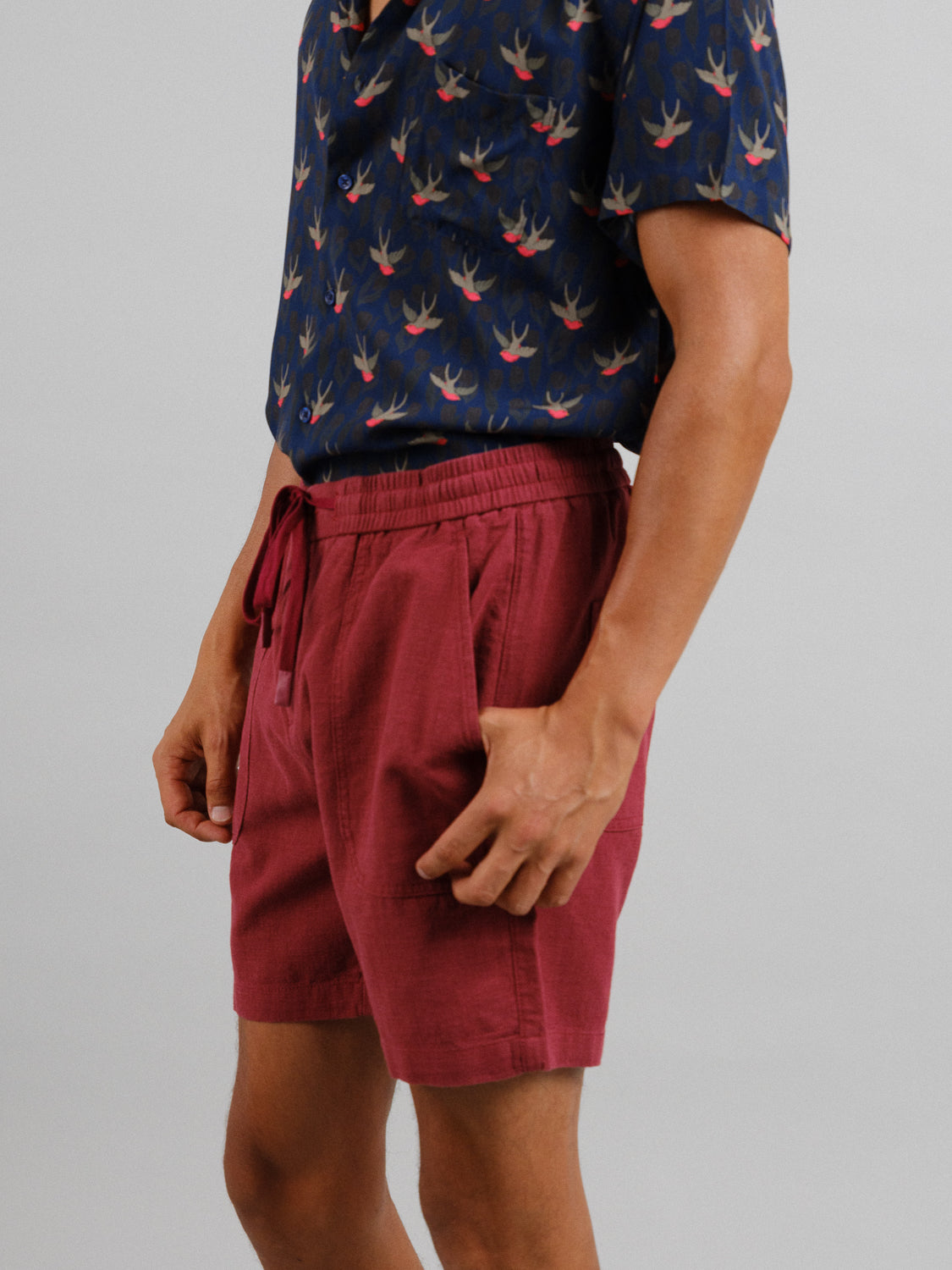 Wearing a navy shirt with a bird pattern, someone models maroon Phi Phi Linen Blend Drawstring Shorts by ID Menswear against a light gray backdrop. Their left hand rests in the shorts pocket, highlighting the casual style and elastic waistband.