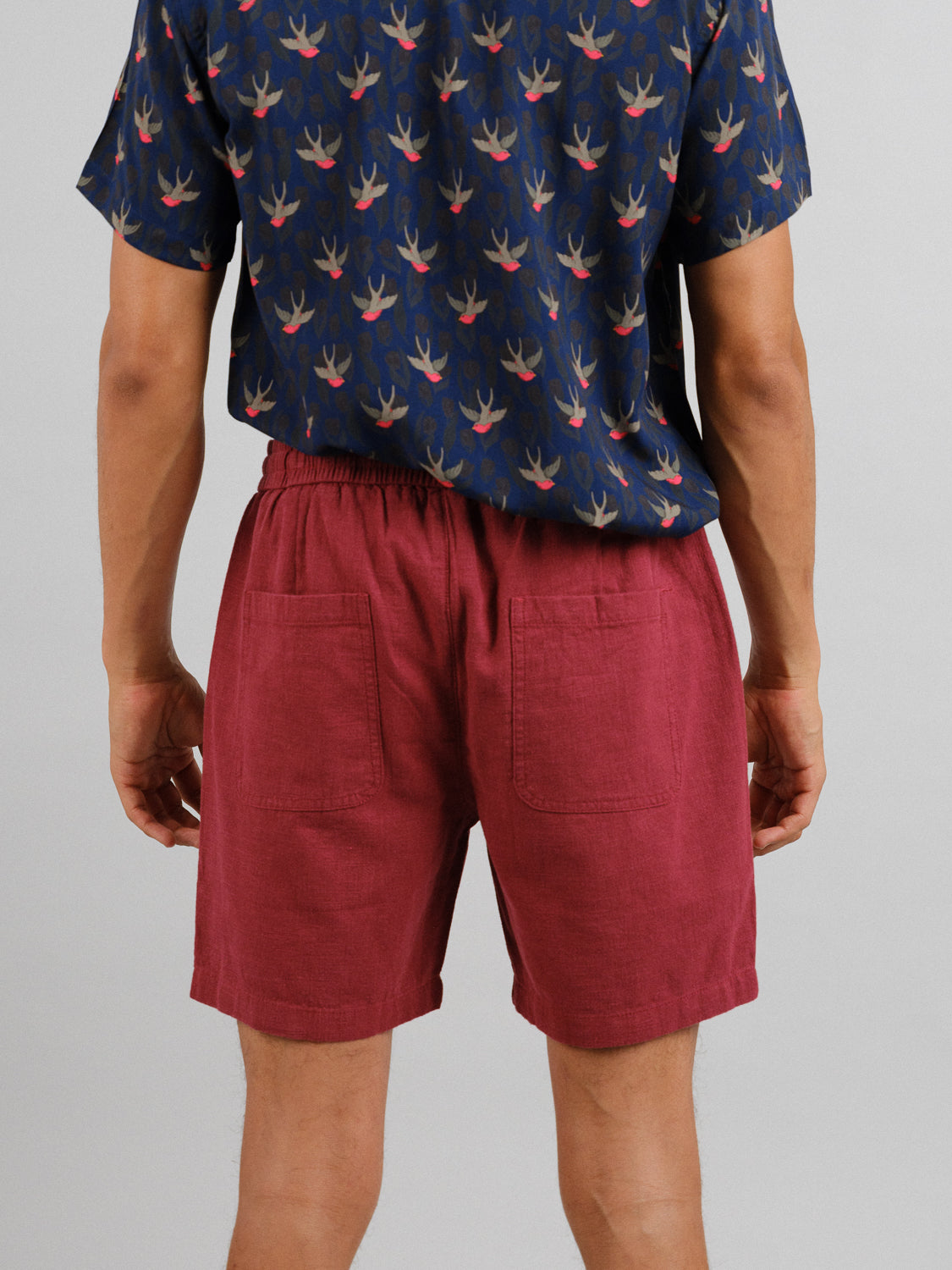 Someone faces away wearing ID Menswears navy bird-patterned shirt and vibrant red Phi Phi Linen Blend Drawstring Shorts, showcasing the 6 inseam and back pockets against a plain light gray background.