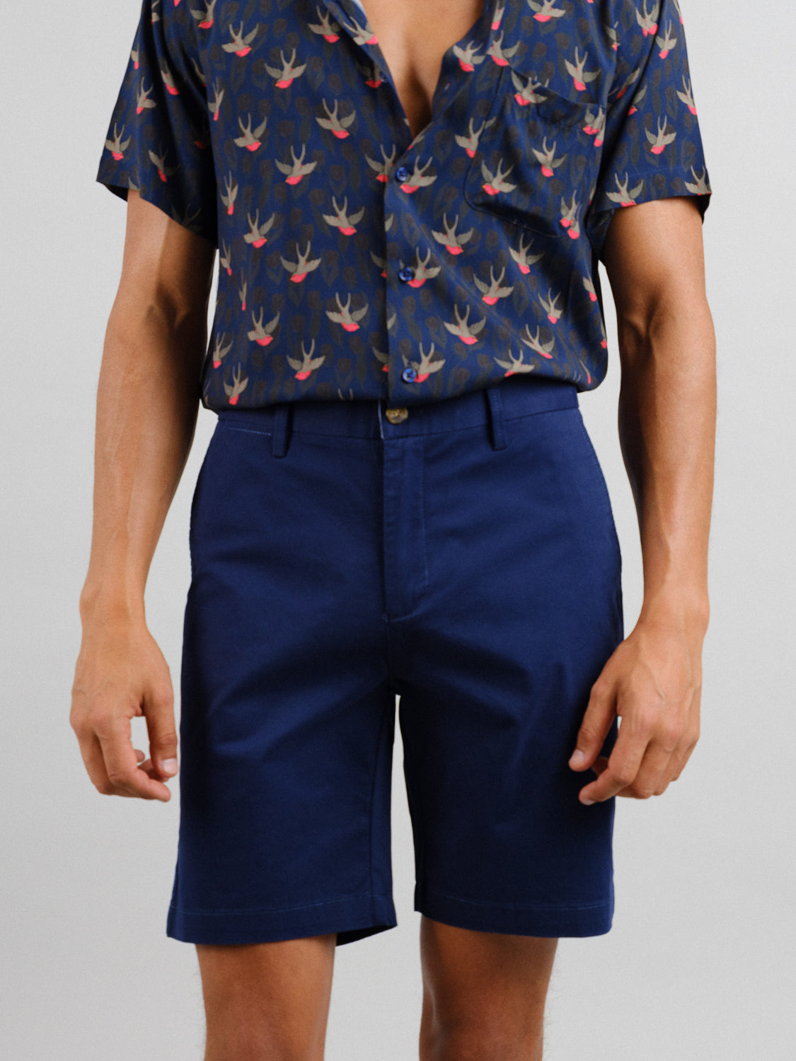 A person stands against a plain background, wearing ID Menswears navy bird-patterned shirt and At Ease Relaxed Chino Shorts in navy. The unbuttoned top of the shirt enhances the outfits relaxed, casual style.