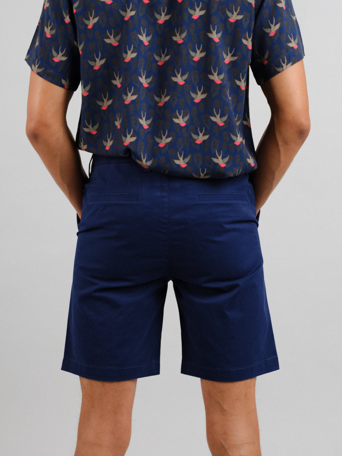 Back view of a person in a navy blue, bird-patterned shirt and At Ease Relaxed Chino Shorts by ID Menswear, hands casually in pockets against a plain gray backdrop.
