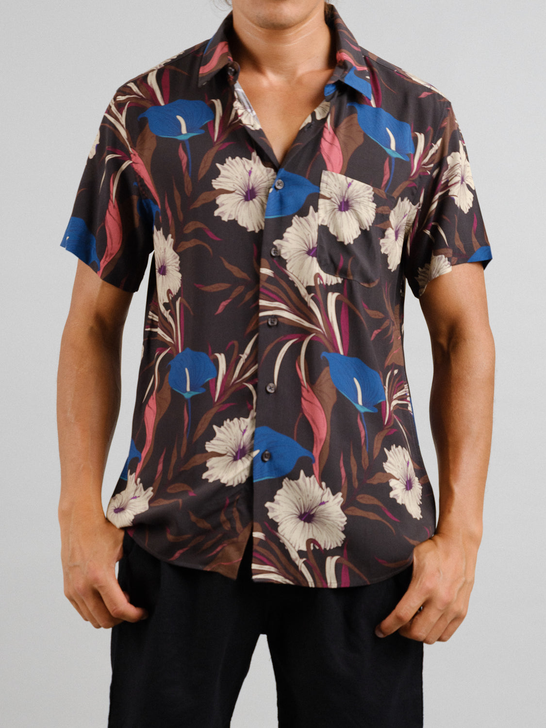 A person wears the Hibiscus Short Sleeve Printed Rayon Shirt by ID Menswear, showcasing vibrant floral prints in blue, white, and burgundy on a dark background. Paired with black pants for warm weather, the look is set against a plain gray backdrop.