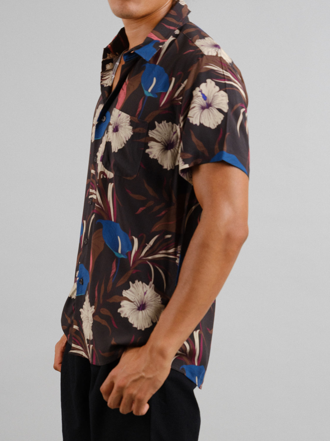 A person dressed in a Hibiscus Short Sleeve Printed Rayon Shirt by ID Menswear, featuring vibrant blue and beige floral patterns, stands against a gray backdrop with black pants. The focus is on the shirts bright design, ideal for warm weather, with their face partially visible.