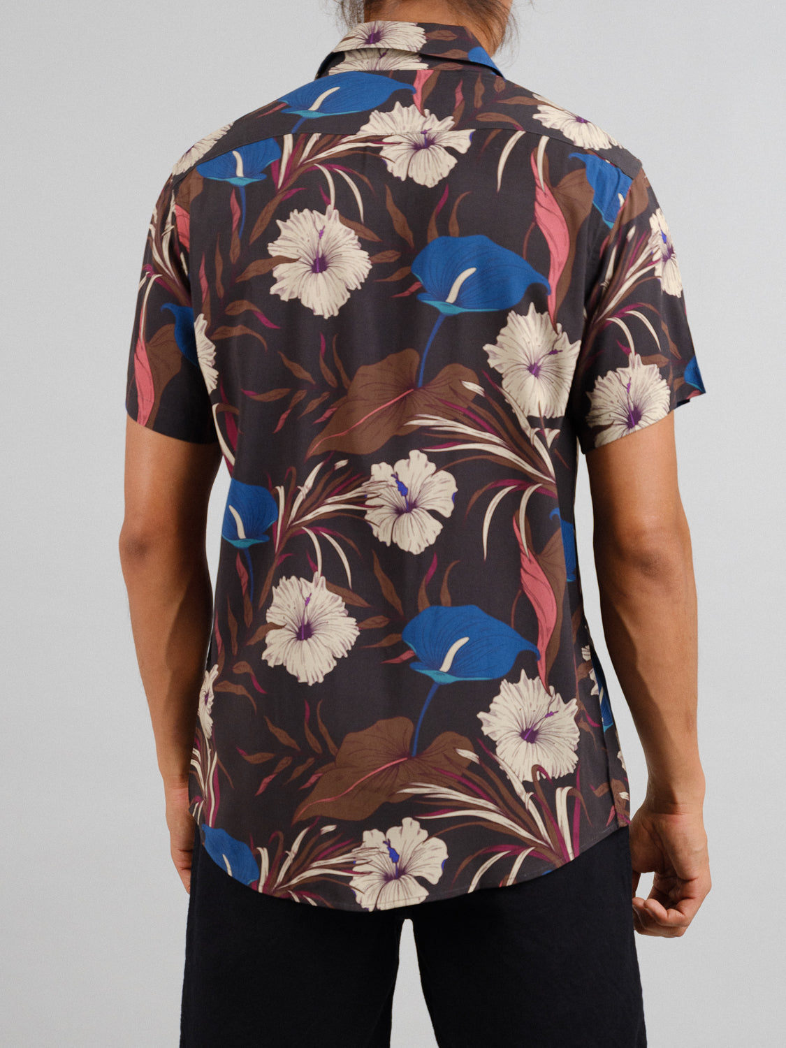 Wearing the Hibiscus Short Sleeve Printed Rayon Shirt by ID Menswear, featuring vibrant floral prints in blue, white, and red on a dark backdrop, a person stands with black pants visible—ideal for warm weather.