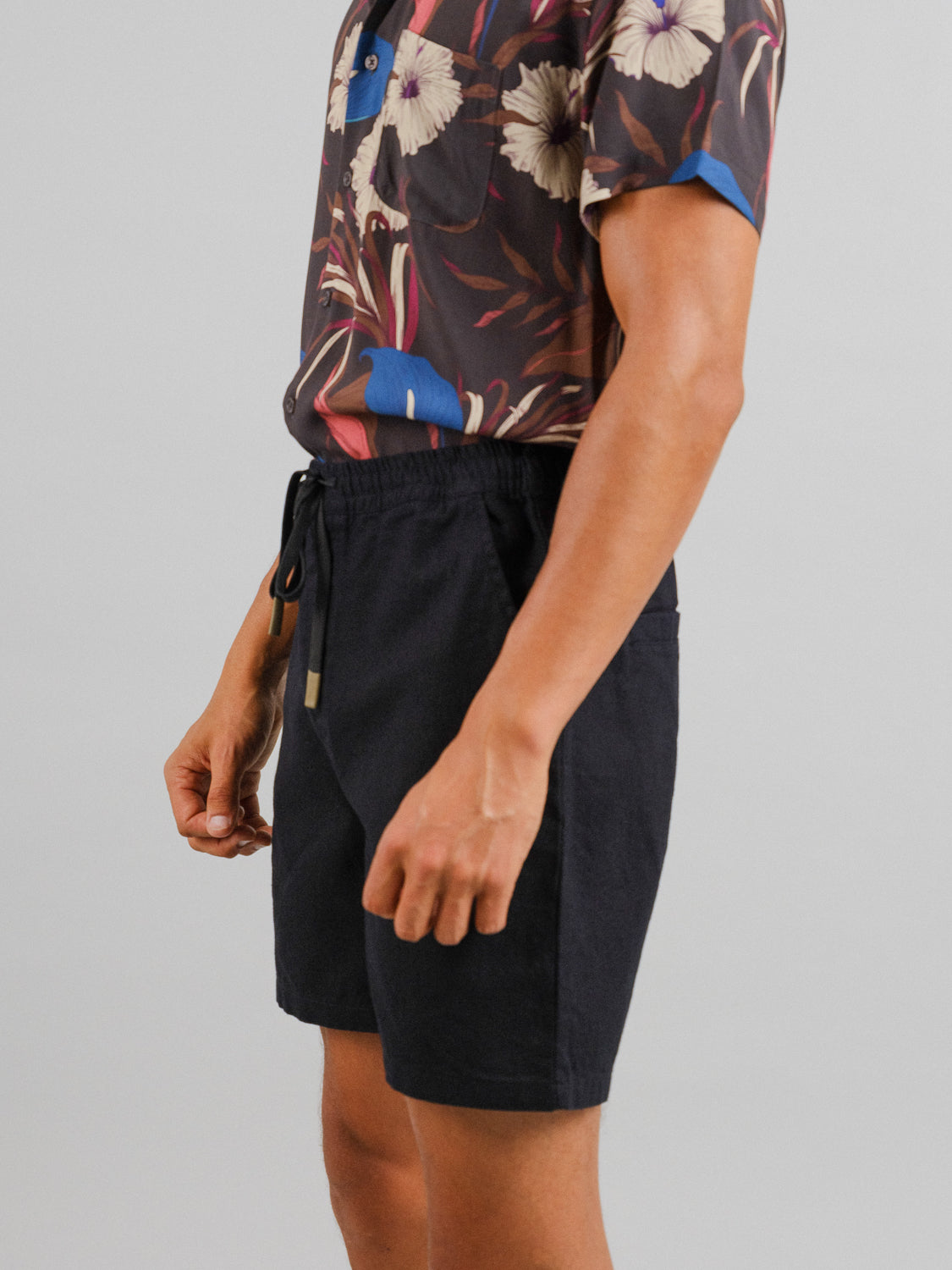 A person wearing a floral shirt and ID Menswears Kotao Linen Blend Drawstring Shorts stands against a light gray background. The shirt features vibrant floral patterns, with focus on the torso and upper legs.