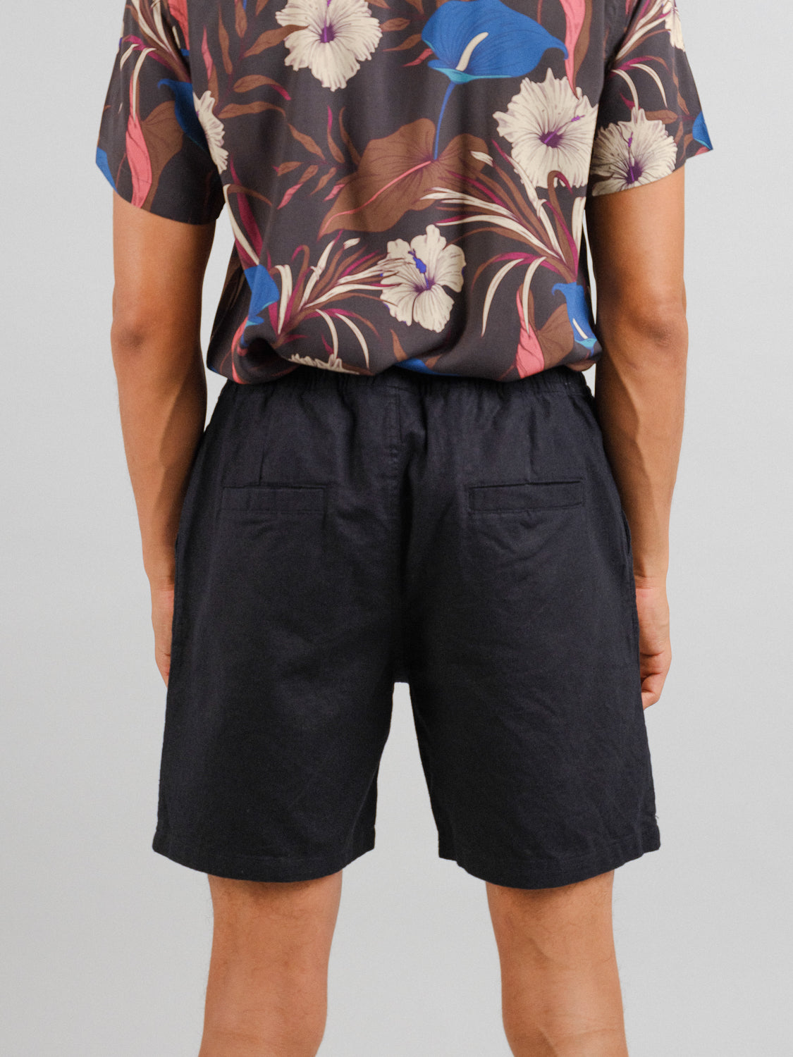 Someone is seen from the back wearing a floral-patterned shirt with ID Menswears Kotao Linen Blend Drawstring Short, featuring large flowers and leaves on a dark background. These black shorts offer a simple yet stylish contrast, perfect for staying cool against the understated backdrop.