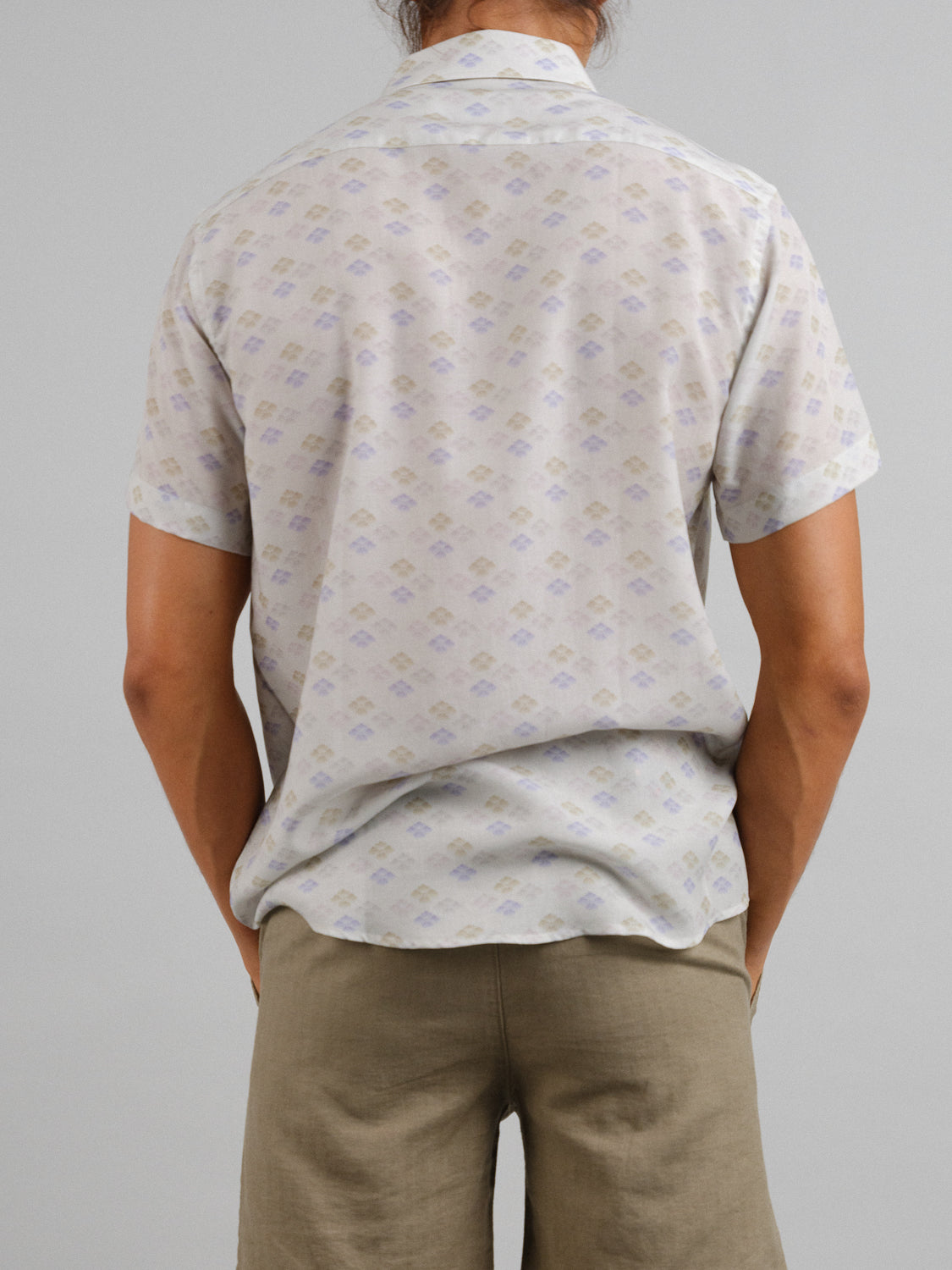 Back view of a person clad in an ID Menswear Hanabi Printed Tencel Linen Shirt paired with beige shorts, epitomizing summer comfort. The shirt has a light background with a geometric pattern. Their long hair is tied back against the plain gray backdrop.