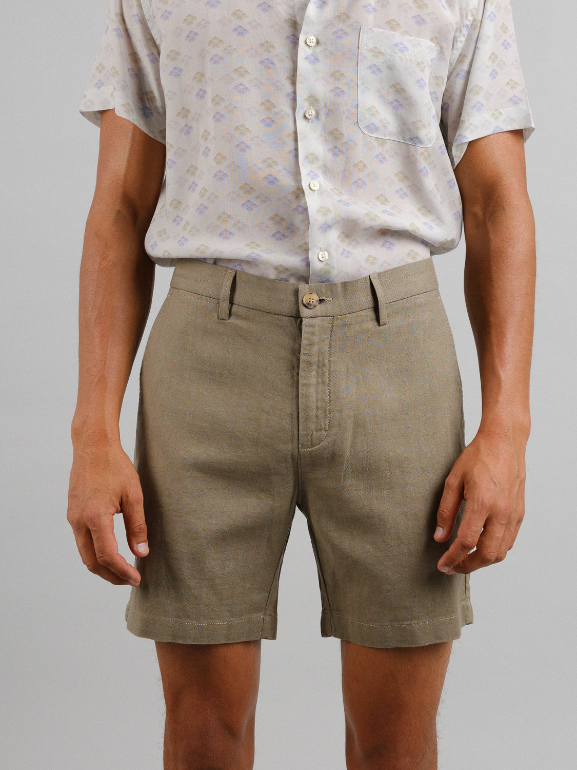 An individual wears ID Menswears Kerala Linen Blend Chino Shorts and a light-patterned, short-sleeve linen-cotton blend shirt with a pocket, tucked in. Their arms rest at their sides against a plain background.