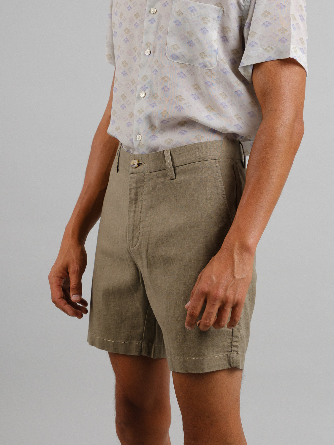 The individual wears a light, patterned short-sleeve shirt made from a linen-cotton blend, paired with beige Kerala Linen Blend Chino Shorts from ID Menswear. Against a plain gray background, the relaxed fit attire is shown from mid-torso to knee with arms casually by their side.