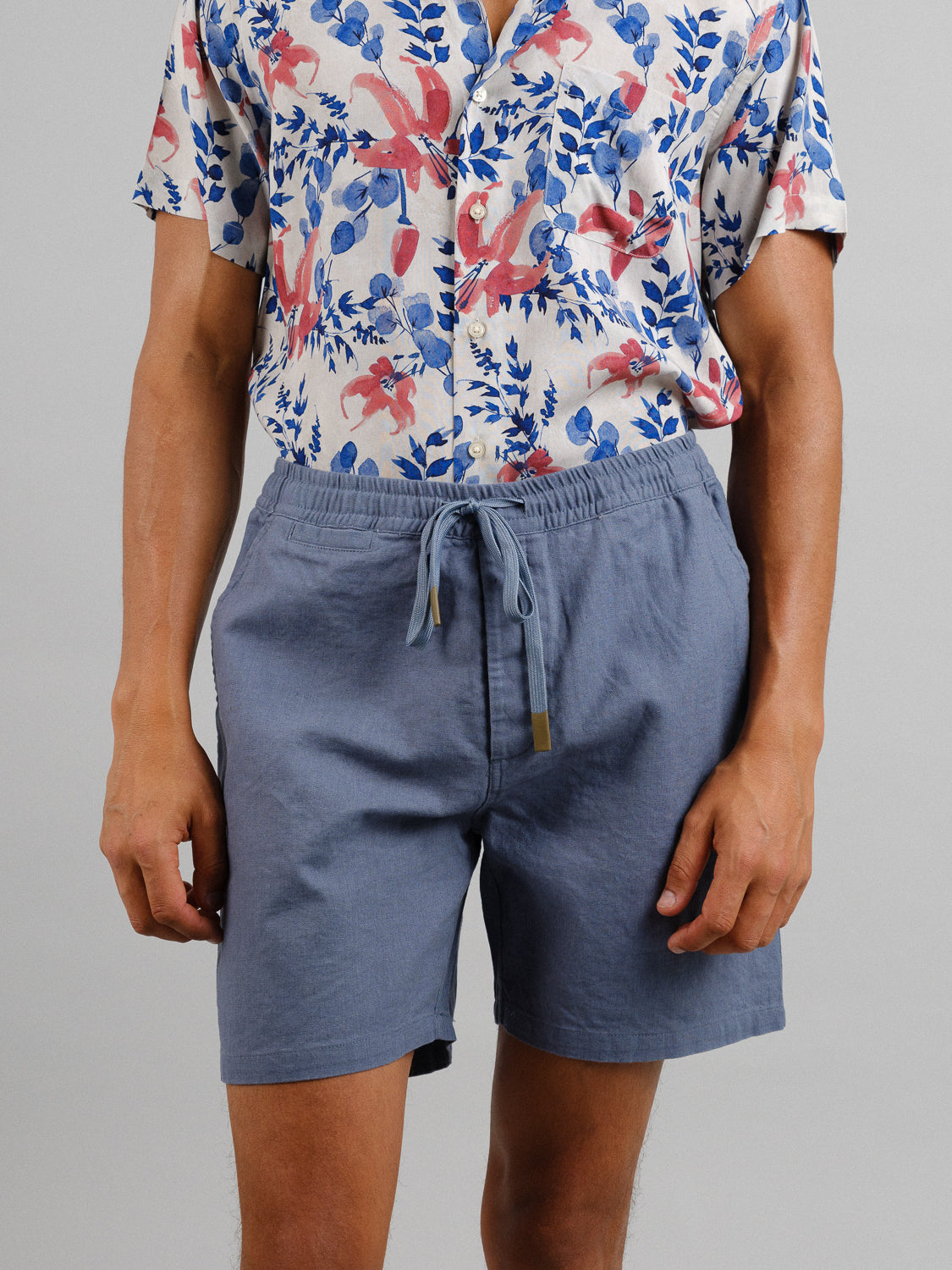 A person in a vibrant floral and leaf-patterned short-sleeve button-up shirt paired with ID Menswears Kotao Linen Blend Drawstring Short exudes a casual, summertime vibe against a plain gray background, perfect for warm days.