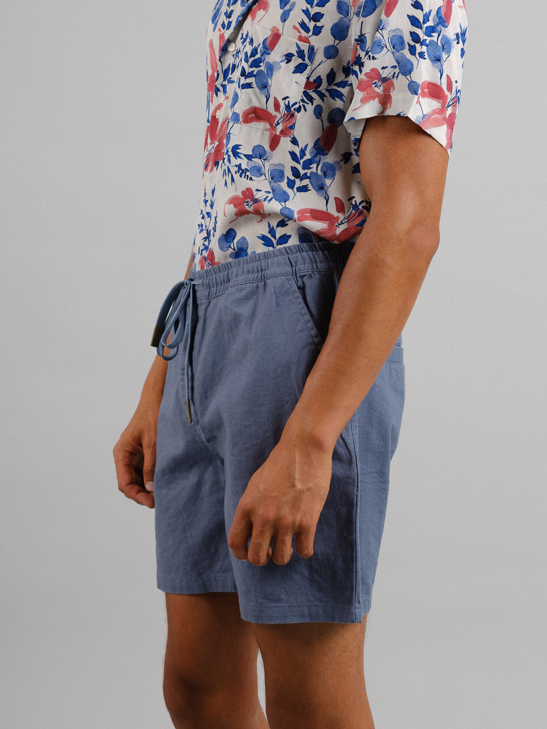 Sporting a floral shirt with red and blue blooms, tucked into ID Menswears Kotao Linen Blend Drawstring Shorts in blue, the individual stands with their left arm relaxed by their side against a plain gray background.