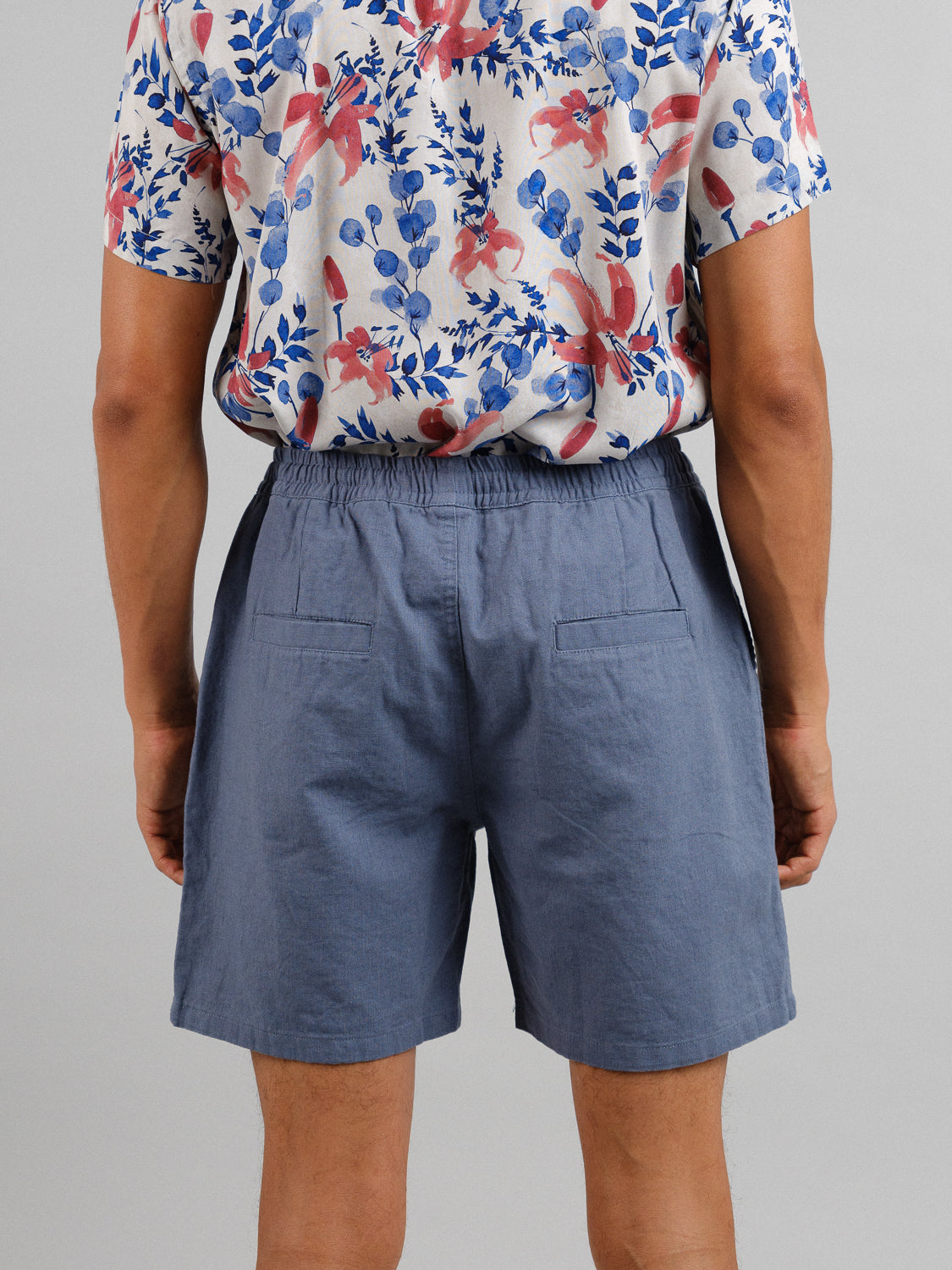 A person wearing a colorful floral shirt featuring red and blue patterns and Kotao Linen Blend Drawstring Shorts from ID Menswear with an elastic waistband stands with their back to the camera against a plain gray background.