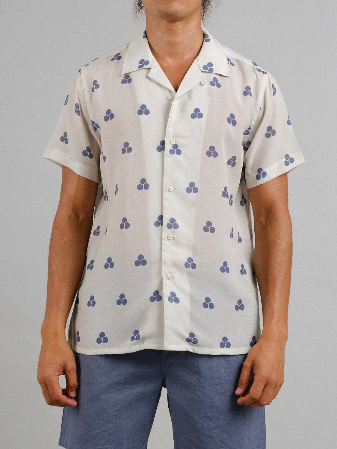 A person wears the Mae Camp Collar Printed Shirt by ID Menswear, known for its breathable comfort and dark blue clover pattern on light fabric. Paired with blue shorts, the ensemble stands out against a simple gray backdrop.