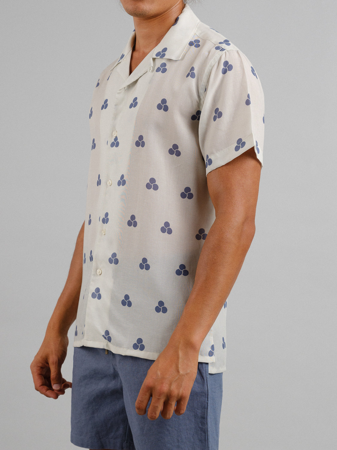 Against a plain gray background, a person exudes effortless style wearing the Mae Camp Collar Printed Shirt by ID Menswear—a light-colored shirt with a dark blue clover pattern, crafted from a breathable tencel-linen blend—paired with blue shorts.