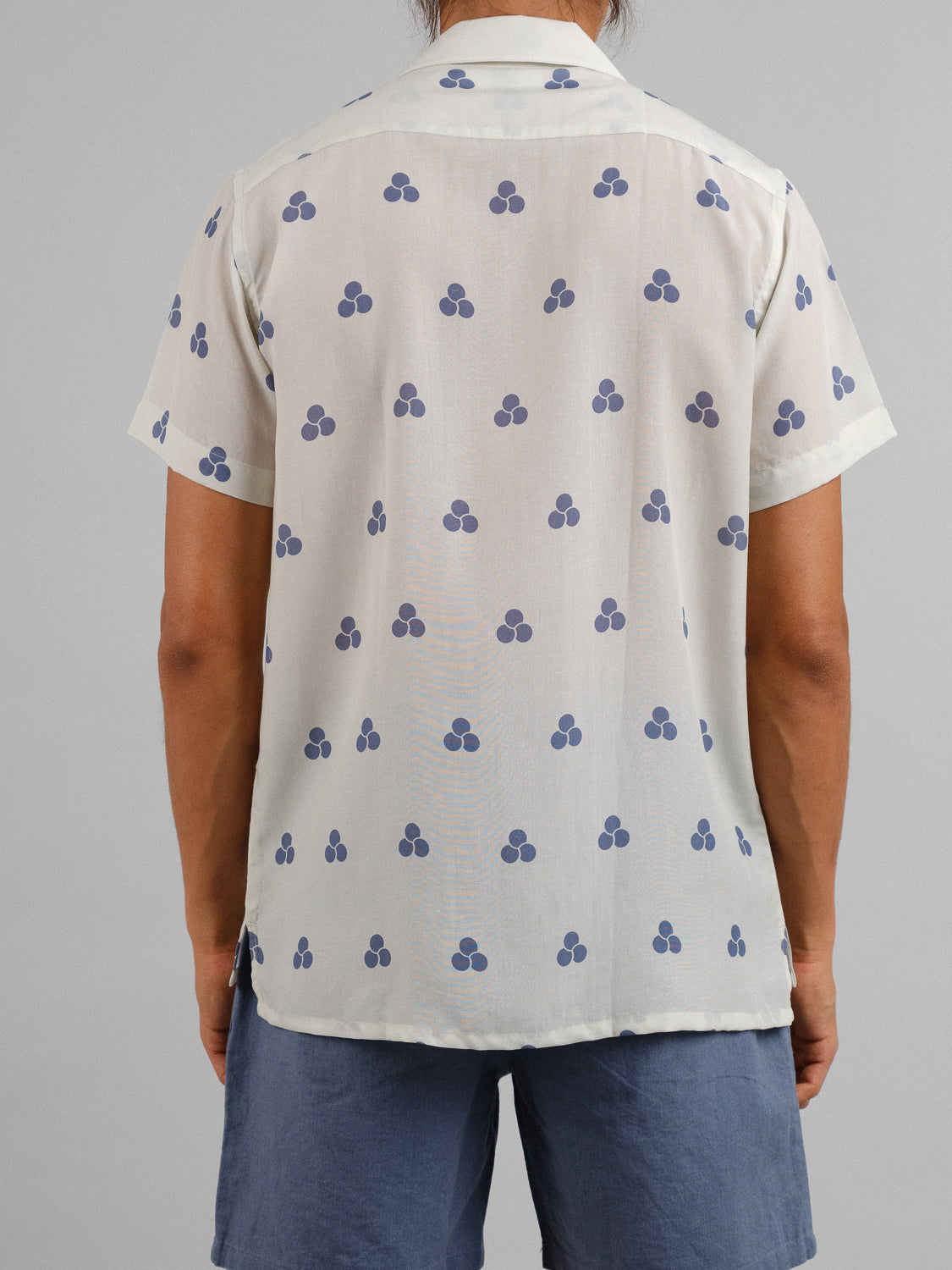 From the back, a person is seen wearing the Mae Camp Collar Printed Shirt by ID Menswear. The shirt features blue triple-dot clusters on light cream tencel-linen fabric for breathable comfort, paired effortlessly with blue shorts against a plain gray background.