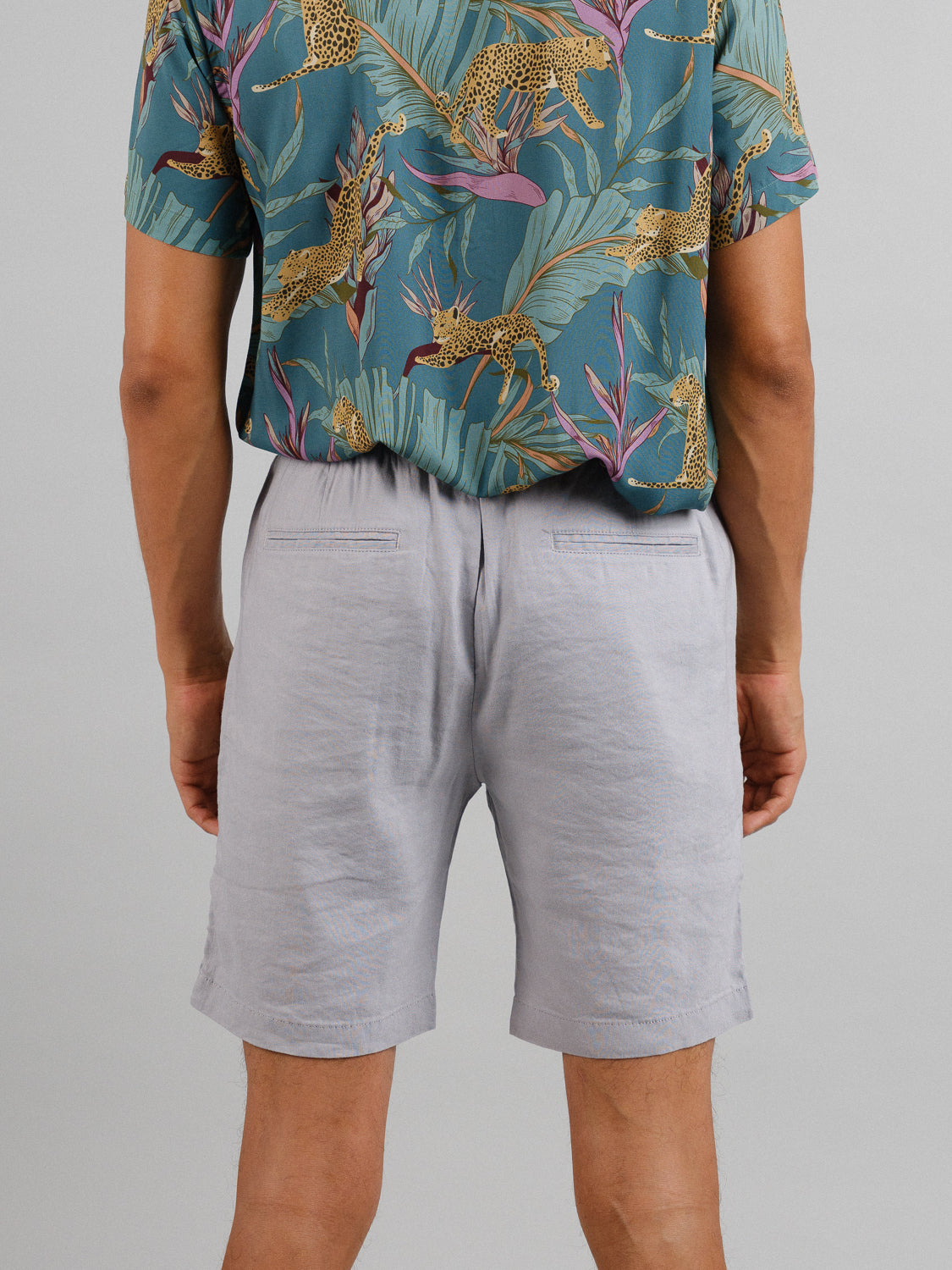 A person faces away from the camera in a green shirt with leopard and leaf patterns, paired with ID Menswears Driggs Linen Blend Short. The light gray shorts are ideal for warm weather, set against a plain, light-colored background.
