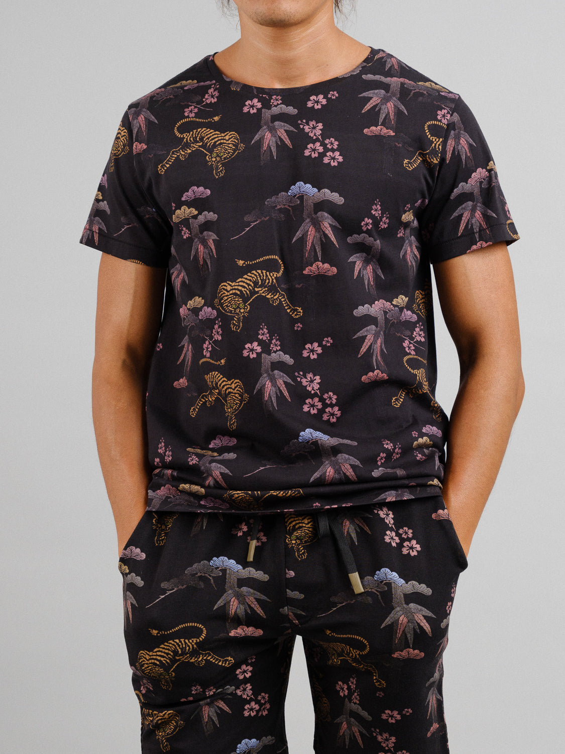 A person wears a Bali Vintage Japanese Print Tee by ID Menswear and matching pants, featuring a vintage tiger, plant, and flower design in muted colors on black organic cotton, standing out against the plain gray background.
