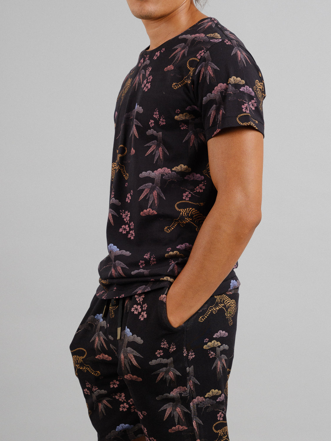 A person in a Bali Vintage Japanese Print Tee by ID Menswear, crafted from organic cotton, with a matching black shirt and pants showcasing tigers, palm leaves, and flowers. One hand rests in their pants pocket against a plain background.