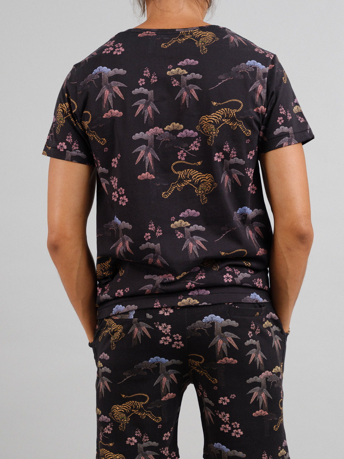 The individual dons ID Menswears Bali Vintage Japanese Print Tee and matching organic cotton pants, adorned with tigers, bamboo, clouds, and pink flowers. With their back to the camera, they embody a refined fusion of elegance and nature.