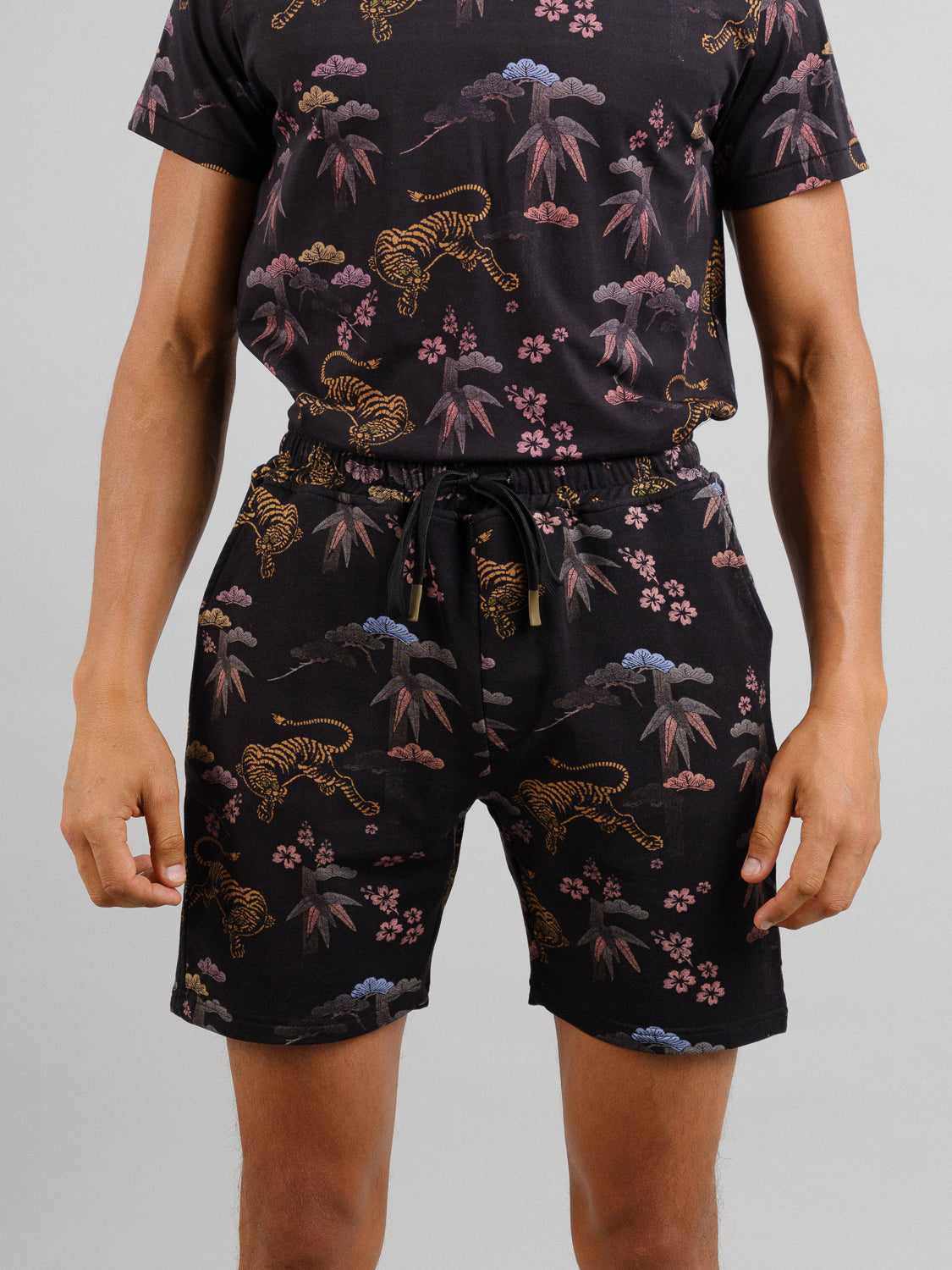A person wears a matching ID Menswear outfit called Samui Vintage Japanese Print Short. The ensemble consists of a short-sleeved shirt and shorts made of organic cotton, featuring nature-themed prints with tigers and plants on black, set against a plain gray background.