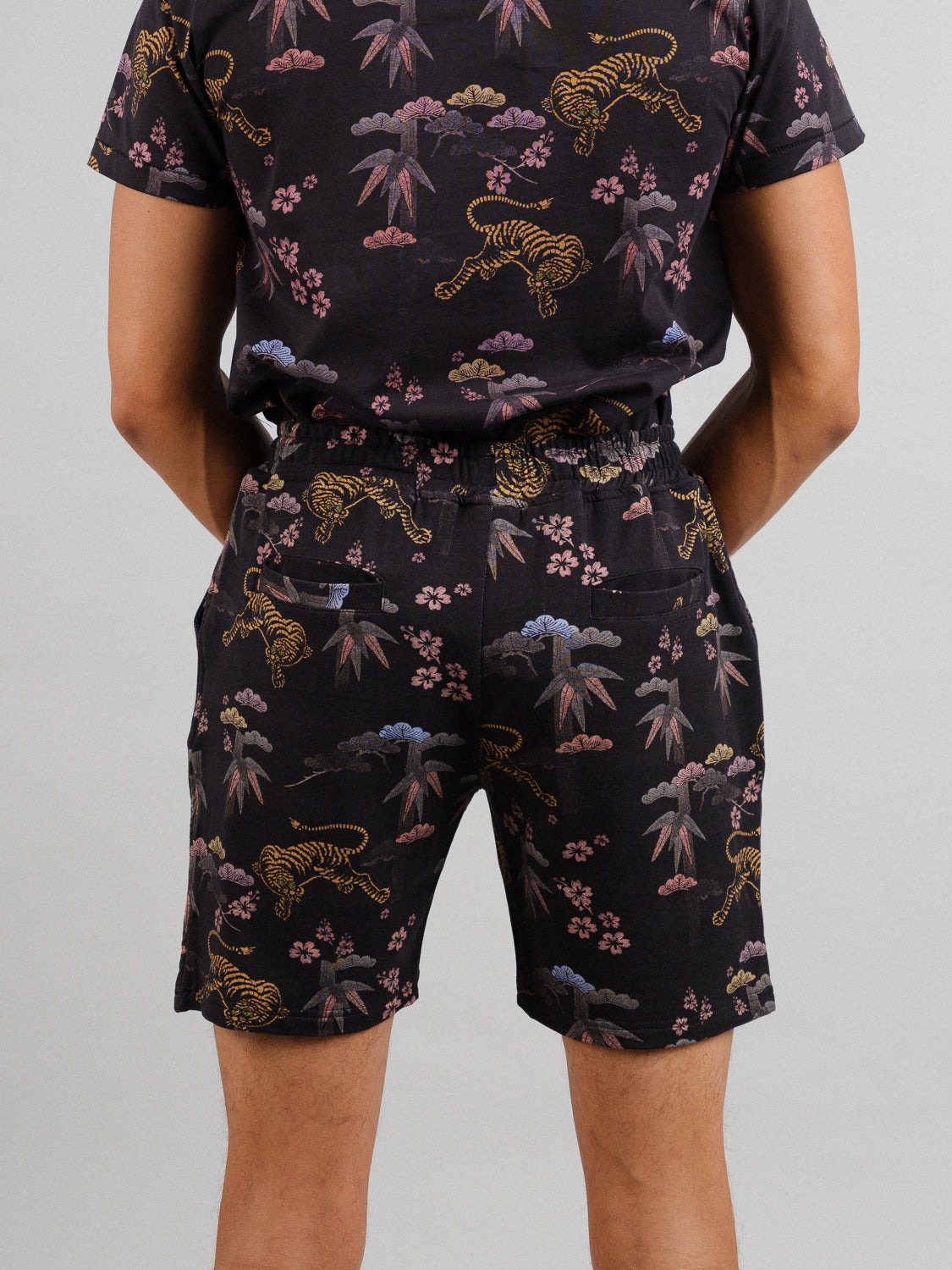 A person wears the Samui Vintage Japanese Print Short from ID Menswear, a matching organic cotton set with a colorful pattern of tigers, bamboo, and clouds on a dark background. The rear view highlights the intricate design reminiscent of vintage Japanese prints.