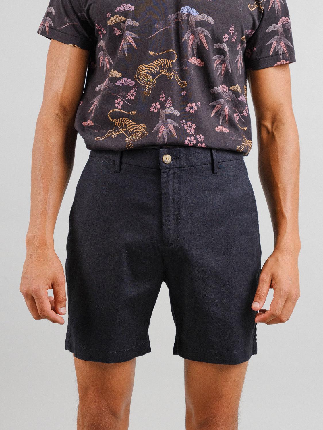 A man stands against a plain background wearing a dark, tropical print shirt with short sleeves and ID Menswears relaxed-fit black Kerala Linen Blend Chino Shorts.