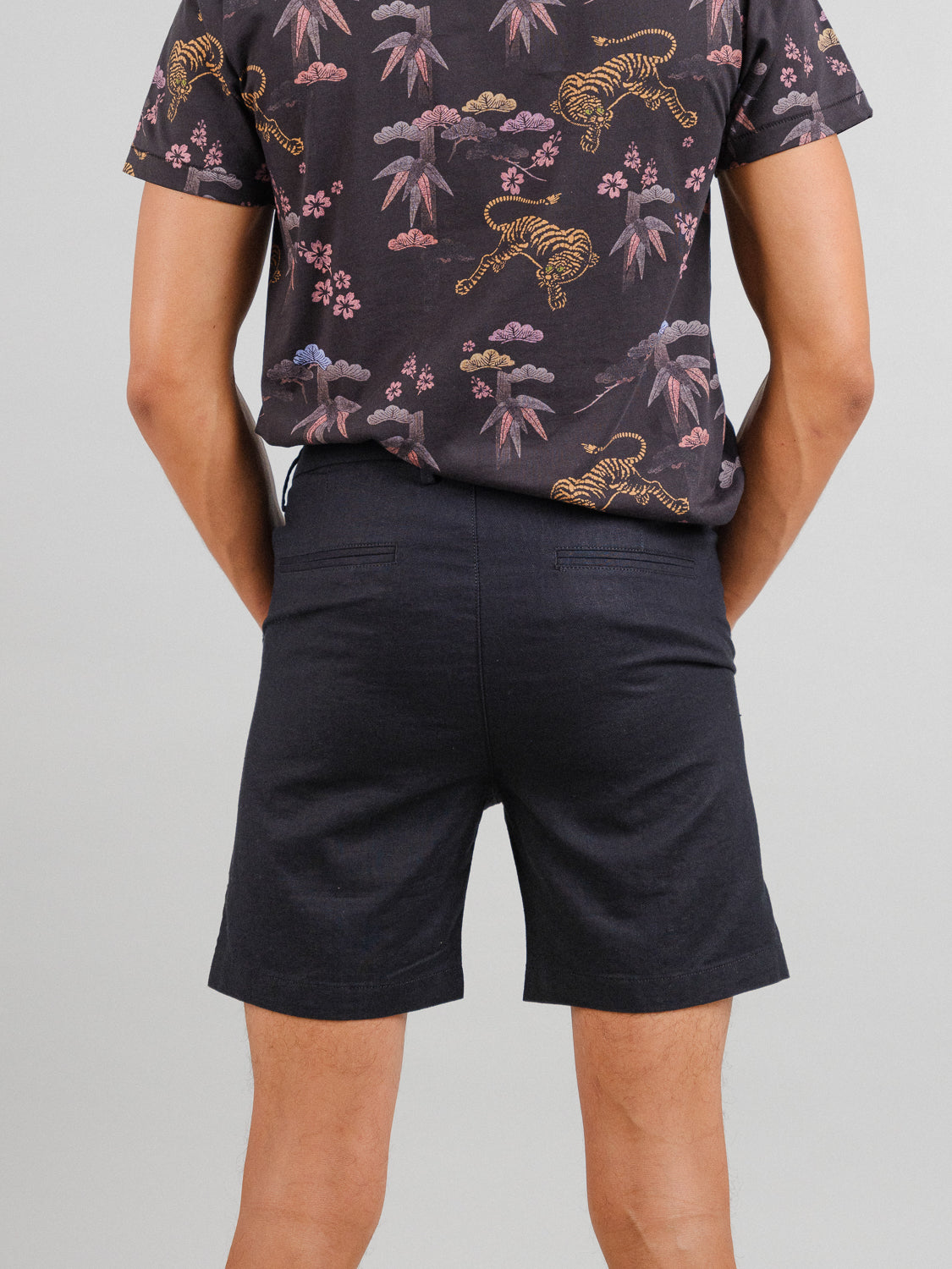 A person in a dark shirt with a vibrant pattern of tigers, mountains, and cherry blossoms faces away from the camera. Theyre wearing relaxed-fit Kerala Linen Blend Chino Short from ID Menswear. The background is plain and light-colored.