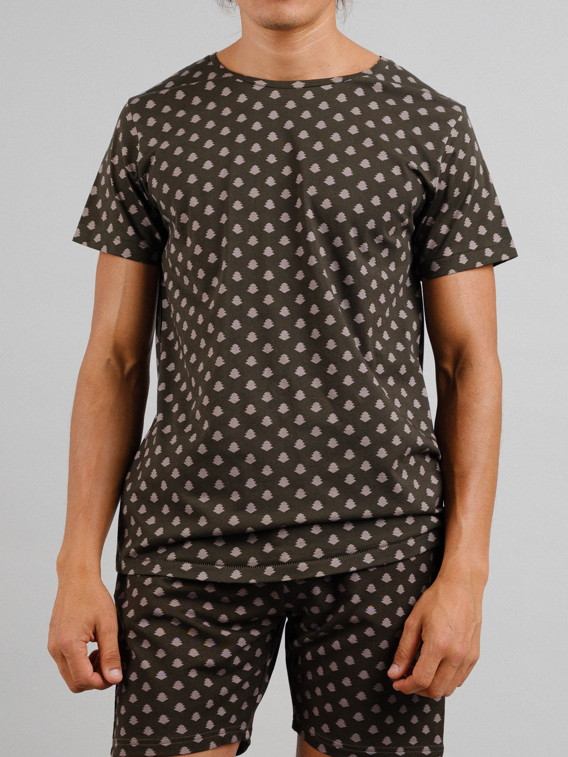 A person in a Bali Vintage Japanese Print Tee from ID Menswear, featuring a matching shirt and shorts set in organic cotton with a dark background and light geometric design reminiscent of vintage Japanese prints, stands relaxed against a plain, light gray backdrop.