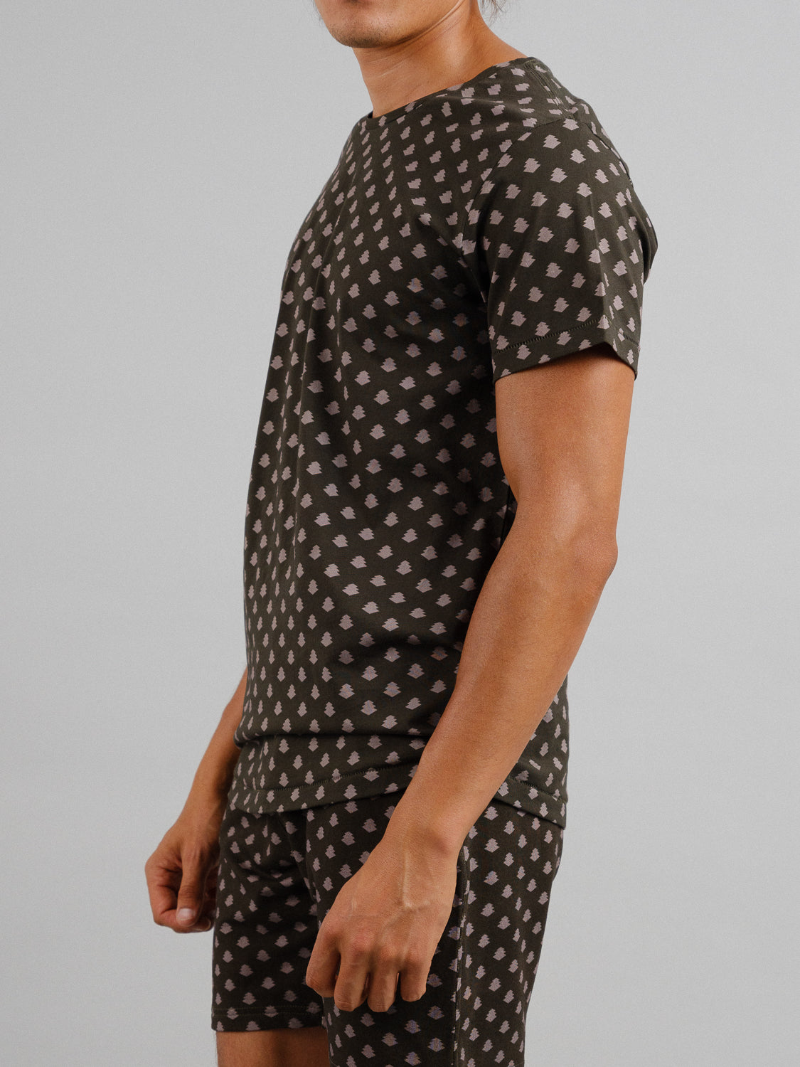 A person stands against a gray backdrop wearing the Bali Vintage Japanese Print Tee and matching shorts from ID Menswear. The outfit, in brown with off-white dots reminiscent of a vintage Japanese pattern, is made from organic cotton. They are photographed in profile.
