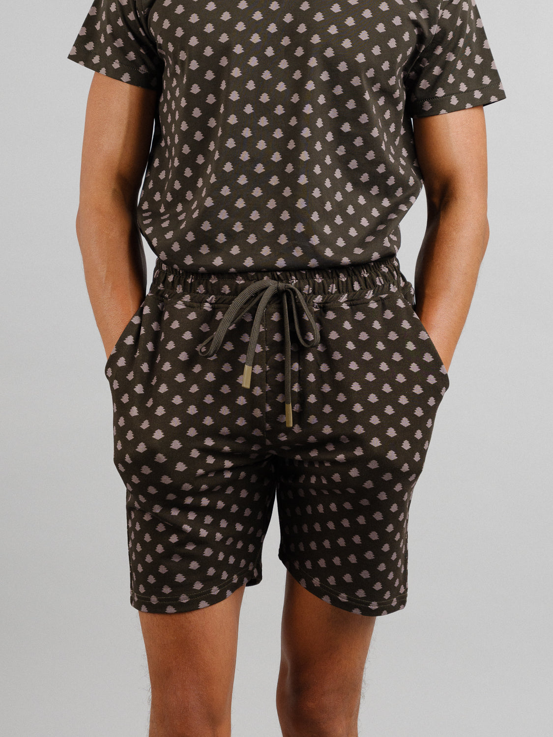 Dressed in a matching set from ID Menswear, the person wears a short-sleeve shirt and Samui Vintage Japanese Print Shorts. Crafted from organic cotton, the outfit features small white diamond patterns on a dark background. They casually stand with hands in pockets against a plain grey backdrop.