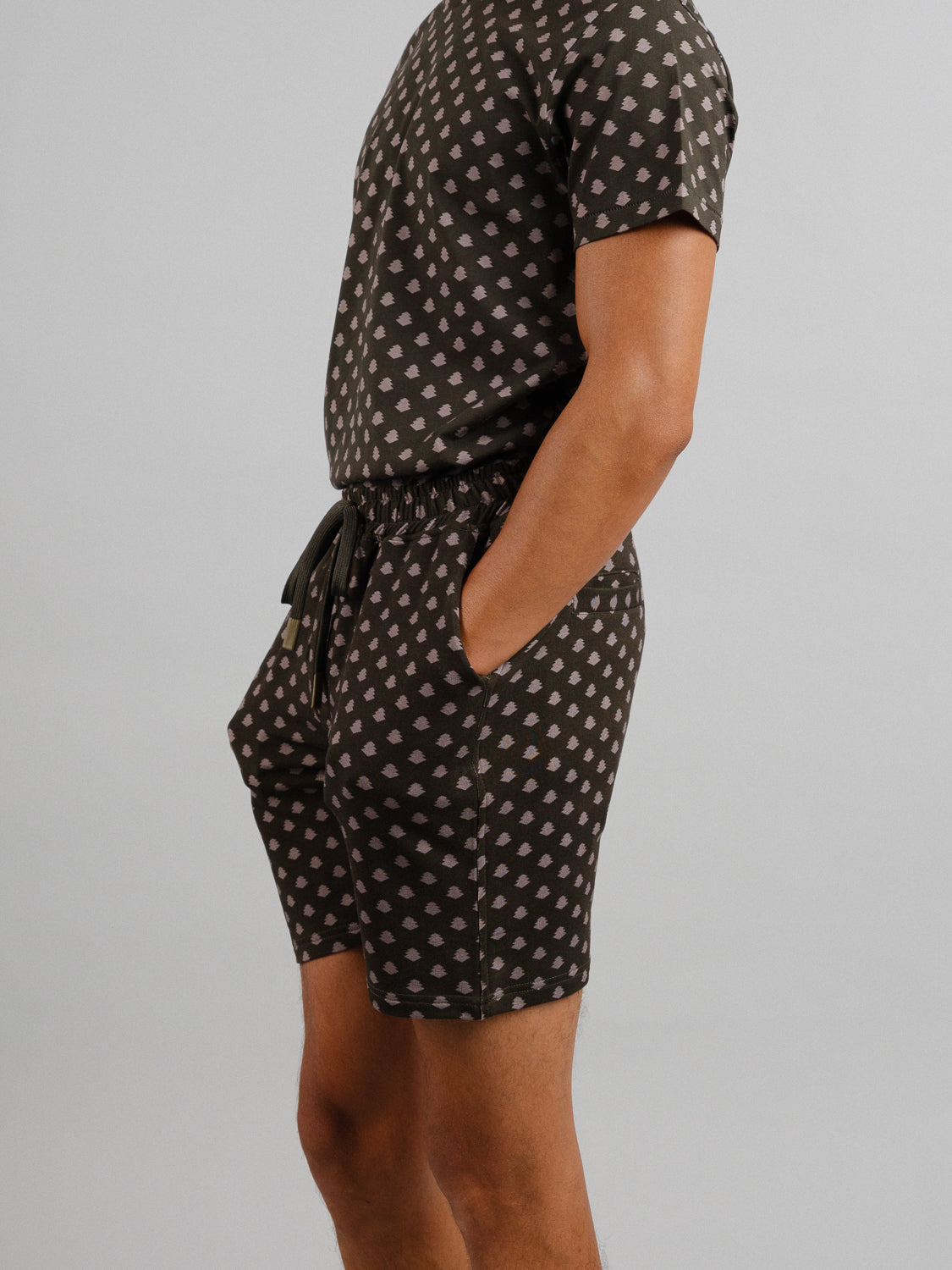 A person wears ID Menswears Samui Vintage Japanese Print Short and a matching short-sleeve shirt, both featuring a light geometric pattern on a dark background. With hands in pockets, they stand slightly turned to the side against a plain backdrop.