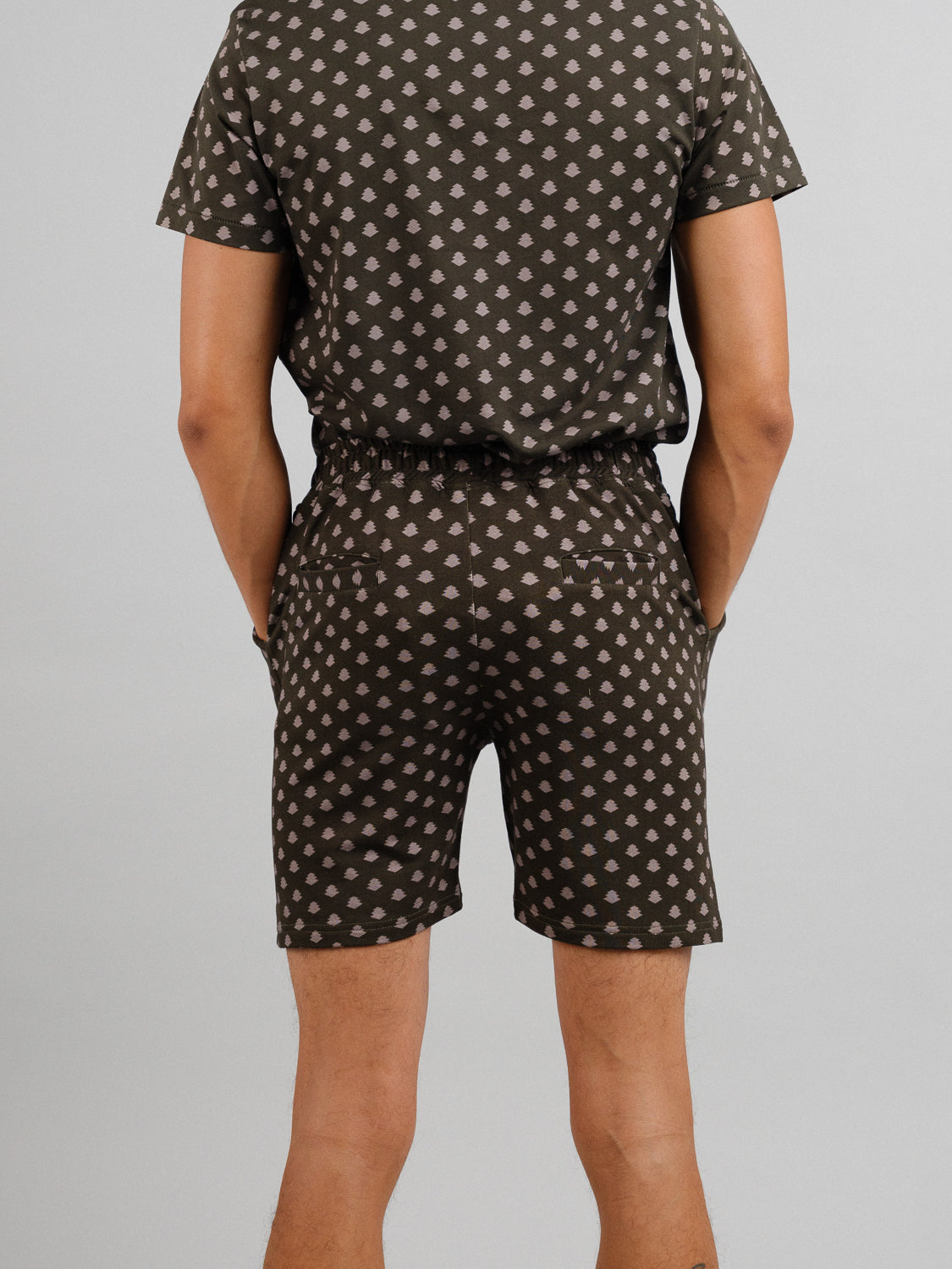 A person is wearing the Samui Vintage Japanese Print Short from ID Menswear. The dark, organic cotton outfit features a short-sleeved shirt and matching shorts with lighter polka dots, evoking a vintage Japanese look as they stand hands behind their back, facing away from the camera.