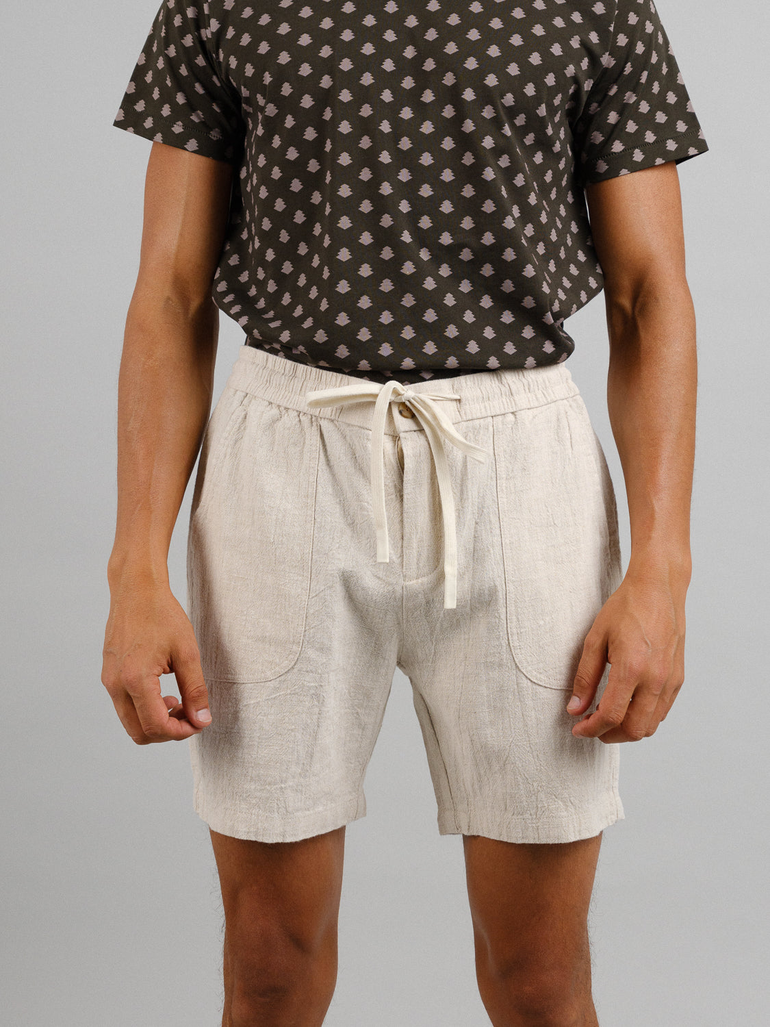 A person in a dark patterned t-shirt effortlessly pairs it with light beige ID Menswear Phi Phi Linen Blend Drawstring Shorts, featuring an elastic waistband and 6 inseam, against a plain gray backdrop. The casual outfit highlights their relaxed pose with hands at their sides.