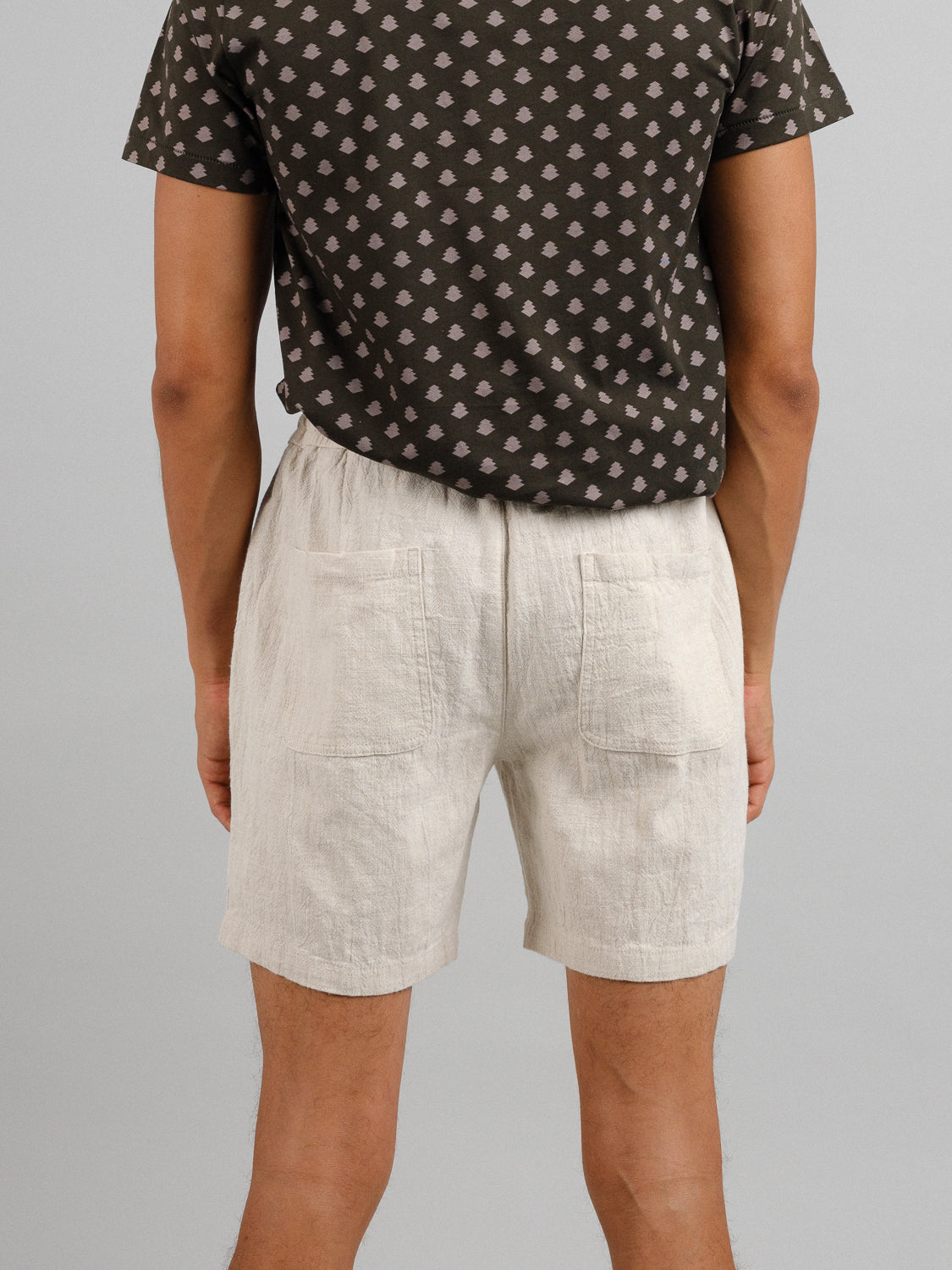 A person faces away from the camera wearing a brown geometric t-shirt and ID Menswears light beige Phi Phi Linen Blend Drawstring Shorts with an elastic waistband and 6 inseam, set against a gray background.
