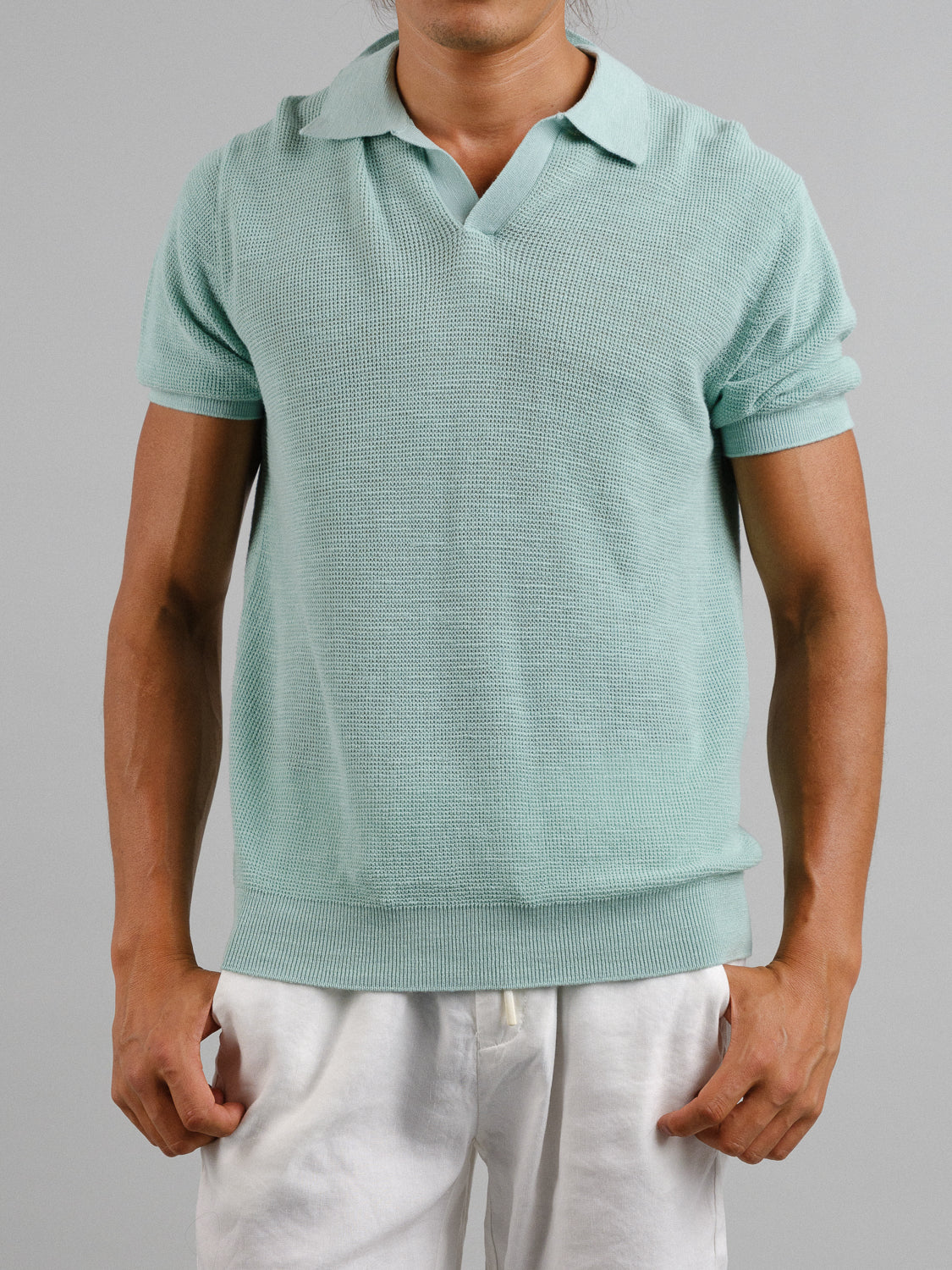 Someone is wearing an Ankara Linen Blend Waffle Knit Polo from ID Menswear, a light green textured short-sleeve sweater ideal for warm weather. Its paired with white pants, and they stand against a plain gray background. The lightweight, breathable fabric offers comfort and style on sunny days.