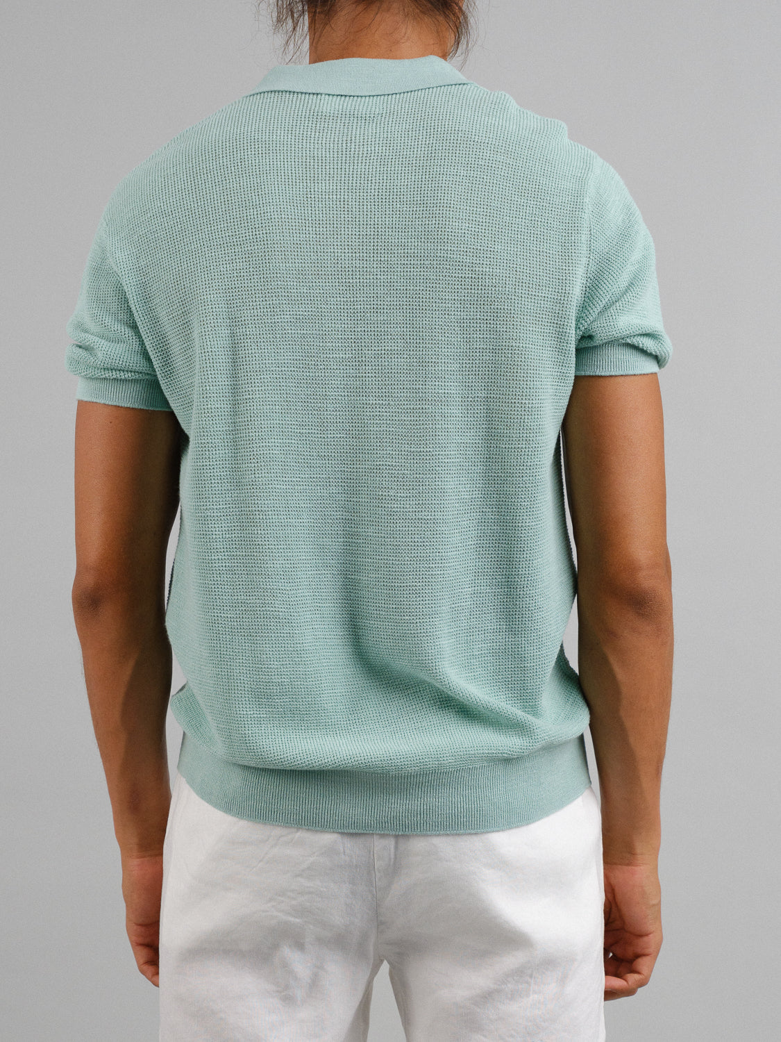 Rear view of a person with long hair, wearing an Ankara Linen Blend Waffle Knit Polo by ID Menswear in light green and white pants against a plain gray background, ideal for warm weather.