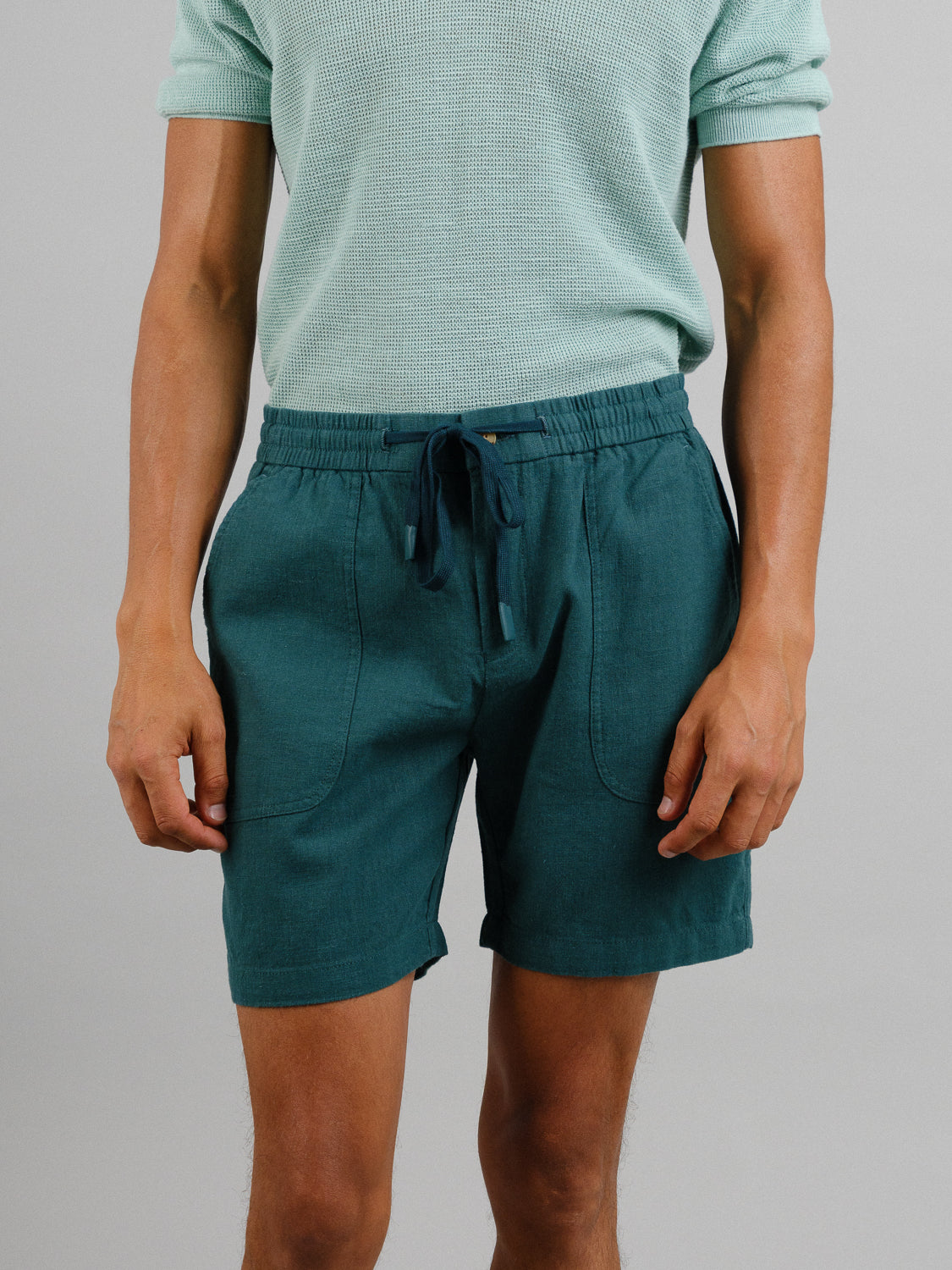 A person in a light green short-sleeve shirt and teal ID Menswears Phi Phi Linen Blend Drawstring Shorts with a 6 inseam, featuring an elastic waistband, stands against a plain gray backdrop, showcasing the outfit from the shoulders down.