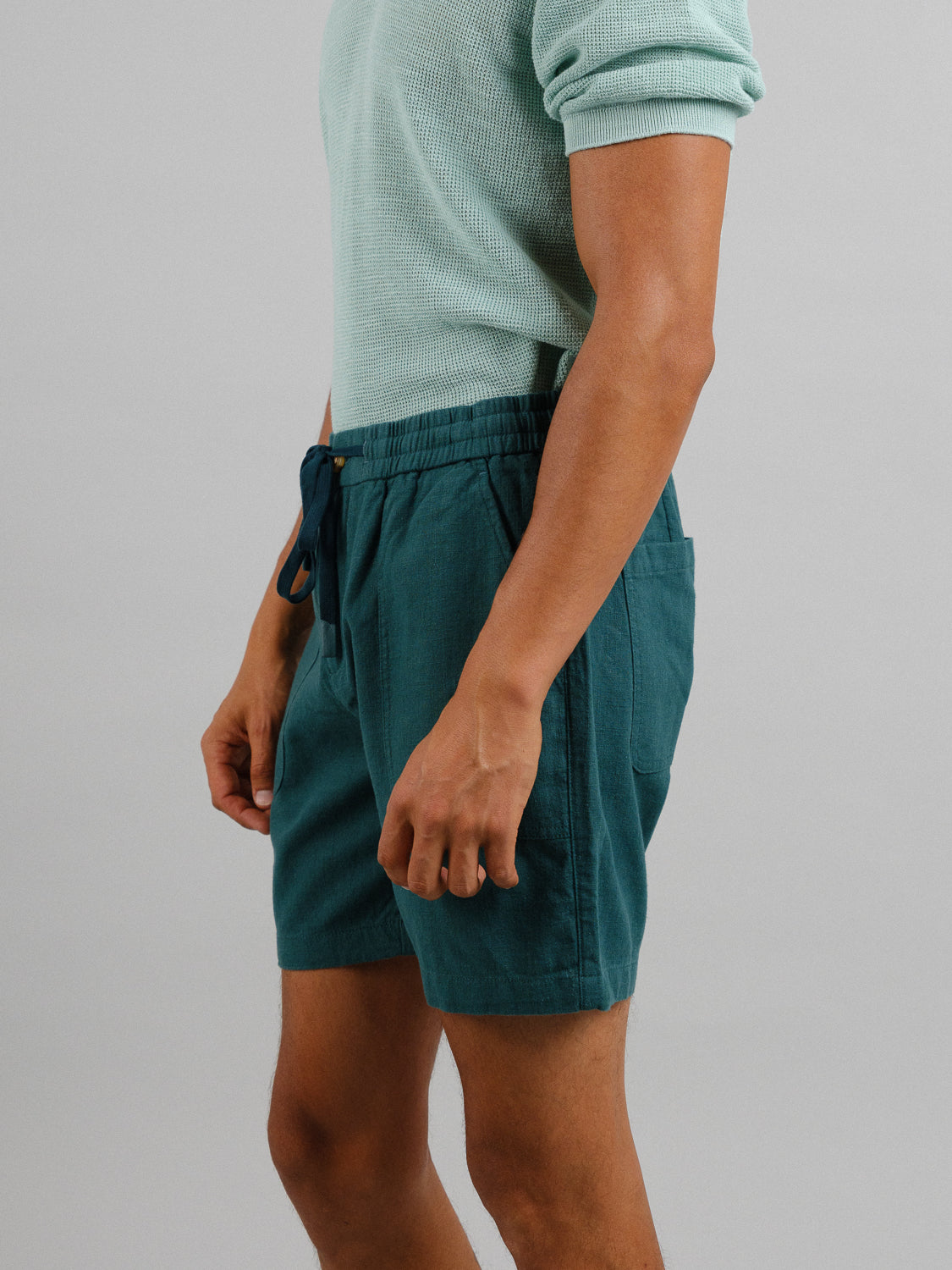 A person wears a textured teal short-sleeve shirt with matching dark teal Phi Phi Linen Blend Drawstring Shorts by ID Menswear against a plain gray background, showcasing the casual outfit with an elastic waistband and relaxed 6 inseam. The image is cropped to show their upper body and thighs.
