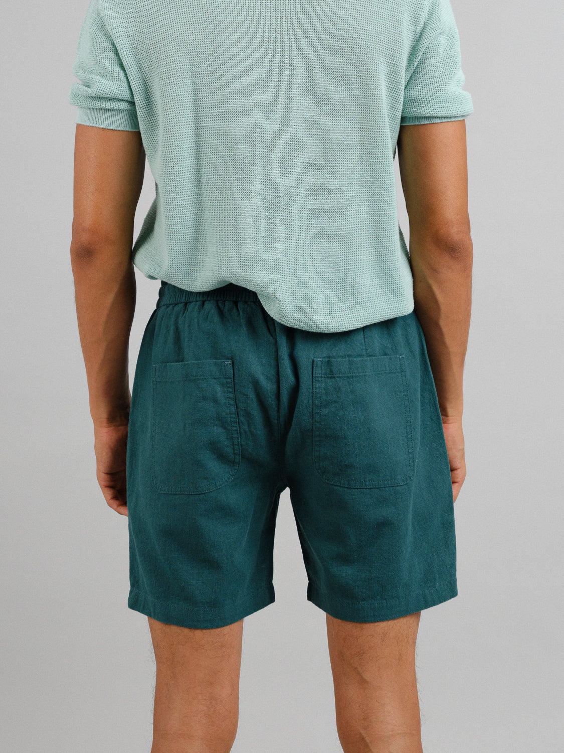 A person seen from behind is wearing a light green textured shirt paired with ID Menswears Phi Phi Linen Blend Drawstring Short in dark teal, featuring an elastic waistband and large back pockets. The simple gray background highlights the 6 inseam, enhancing their casual appeal.