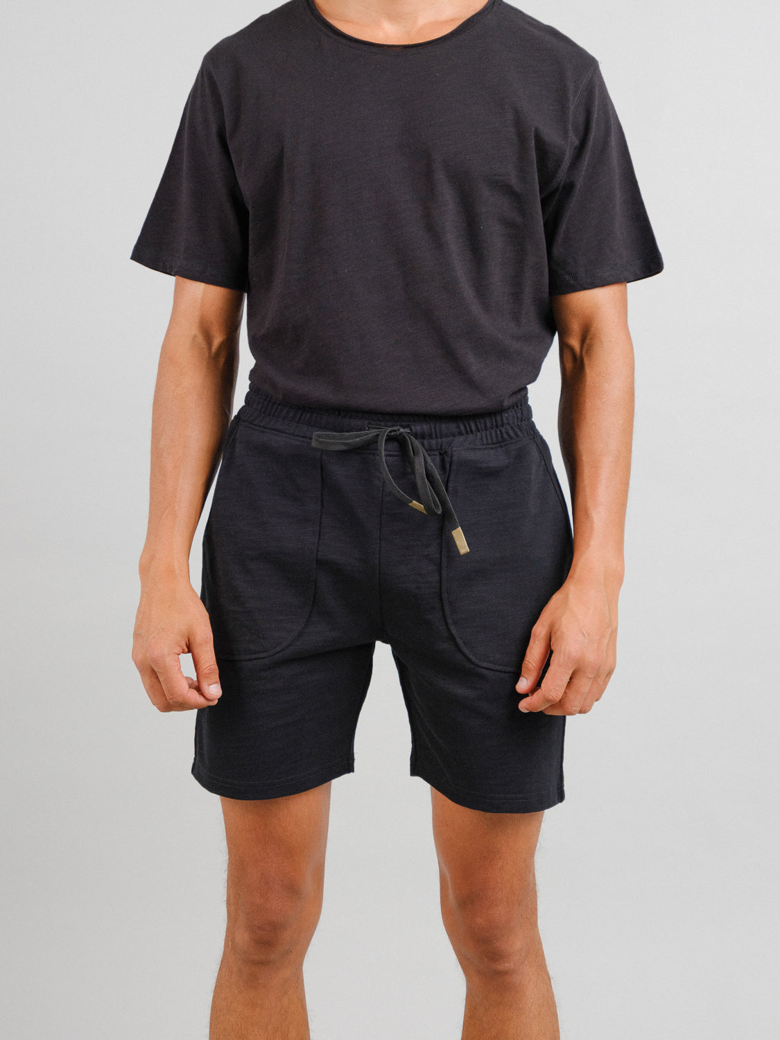 A person wearing a plain black T-shirt and ID Menswears Kahala Slub Cotton Drawstring Short stands against a neutral background. The simple, casual outfit is ideal for everyday wear, with the persons hands relaxed at their sides.