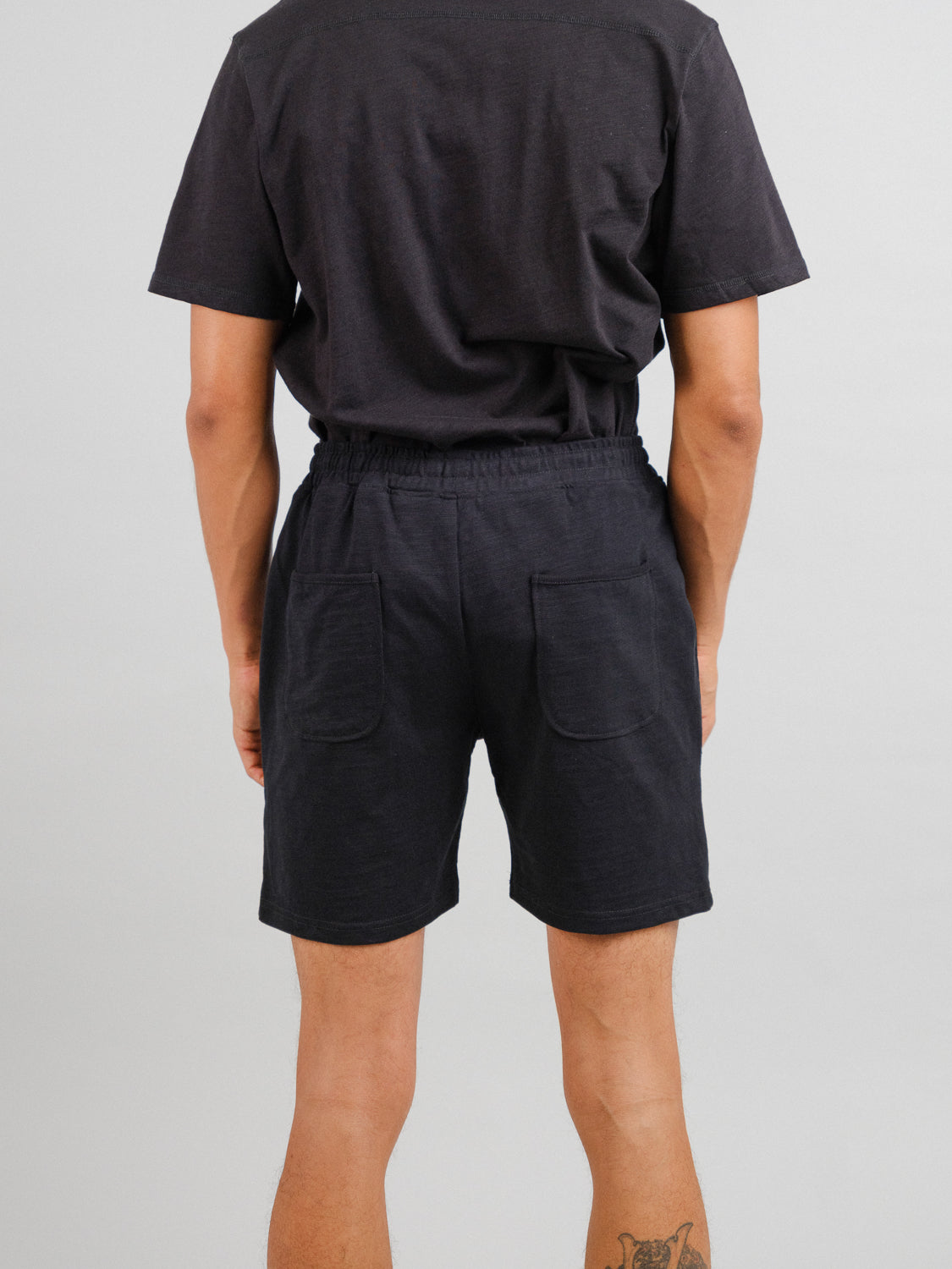 A person wears ID Menswears Kahala Slub Cotton Drawstring Shorts, relaxed fit in black with two back pockets. The backdrop is plain light gray, showcasing a subtle tattoo on their right calf.