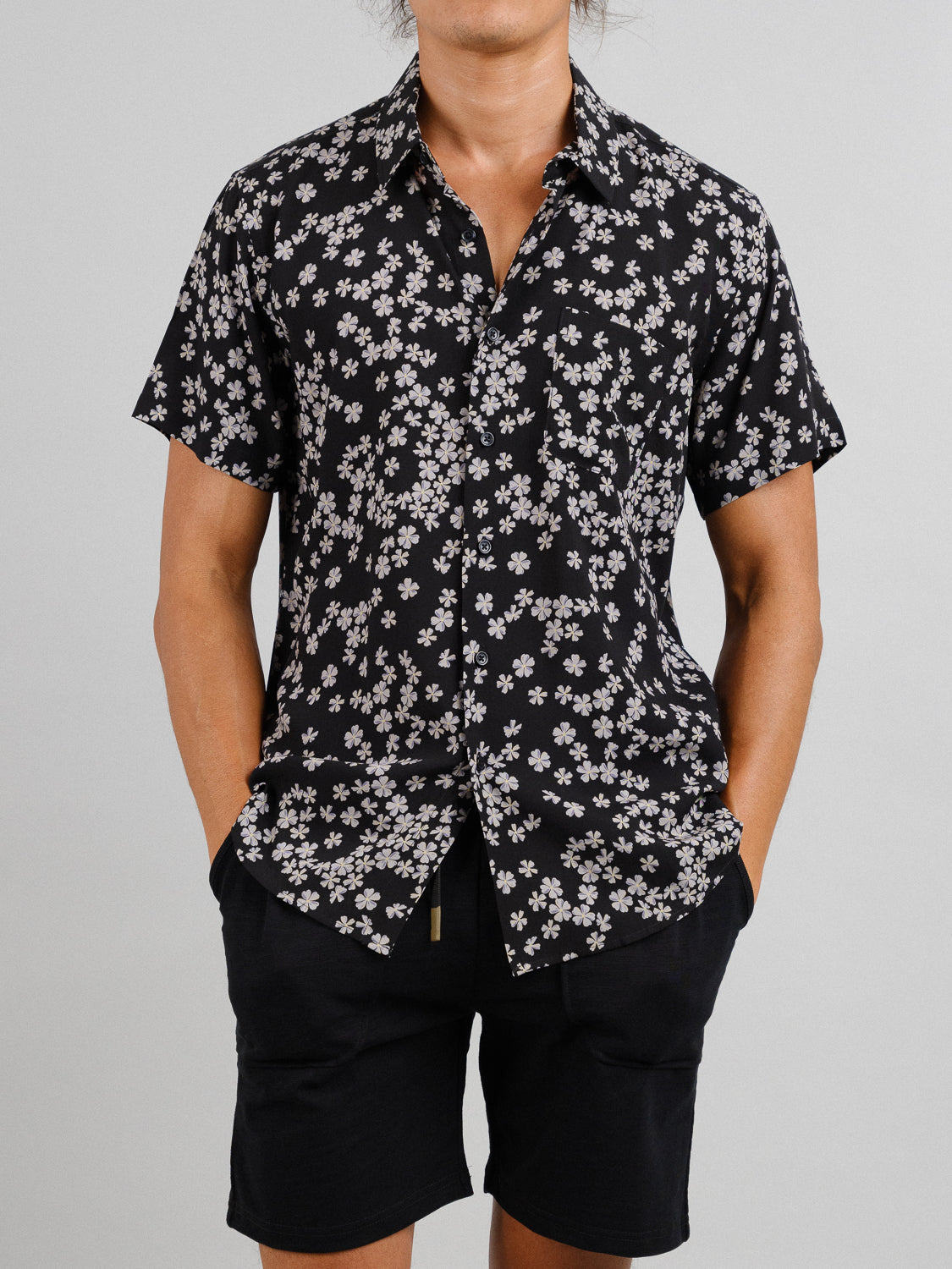 Wearing the Forgetmenot Printed Rayon Shirt by ID Menswear, a black short-sleeve adorned with white flowers, paired with black shorts, the person casually slips their hands into their pockets against a gray background.