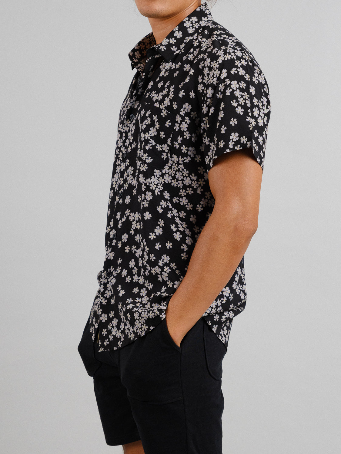 Sporting short black hair, the person wears ID Menswears Forgetmenot Printed Rayon Shirt, a lightweight short-sleeve top with white floral patterns. They pair it with black shorts, hands in pockets, against a plain light gray backdrop.