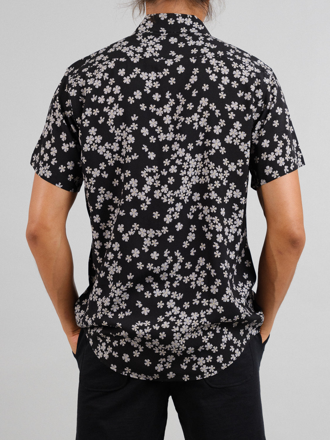 Wearing ID Menswears Forgetmenot Printed Rayon Shirt featuring vibrant white floral designs, a person with long hair stands, hands in pockets of black pants against a plain gray backdrop, facing away from the camera.
