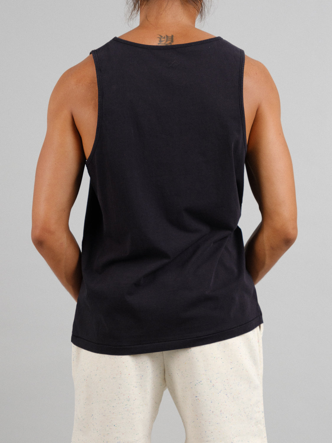 A person with a neck tattoo is seen from the back, wearing an ID Menswear Settler Snow Vintage Wash Tank Top and light-colored pants against a plain backdrop.