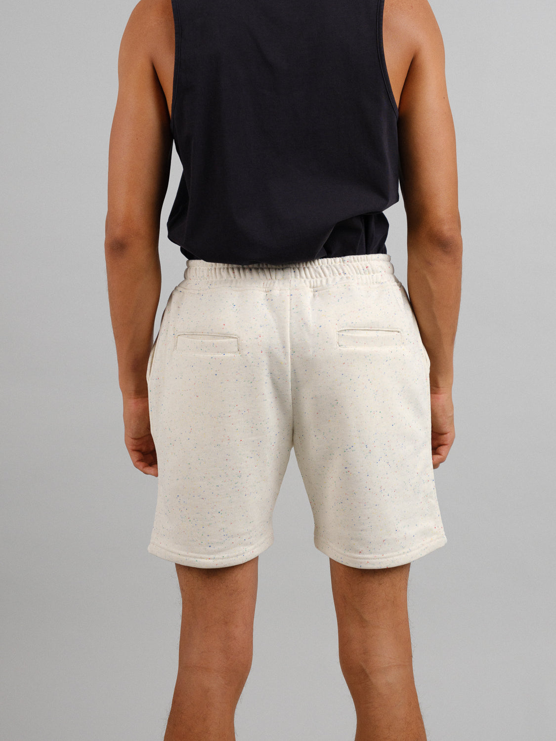 A person stands with legs slightly apart against a gray background, wearing a sleeveless black top and ID Menswears Waikiki Speckled Cotton Shorts, showcasing a fashion-forward style.