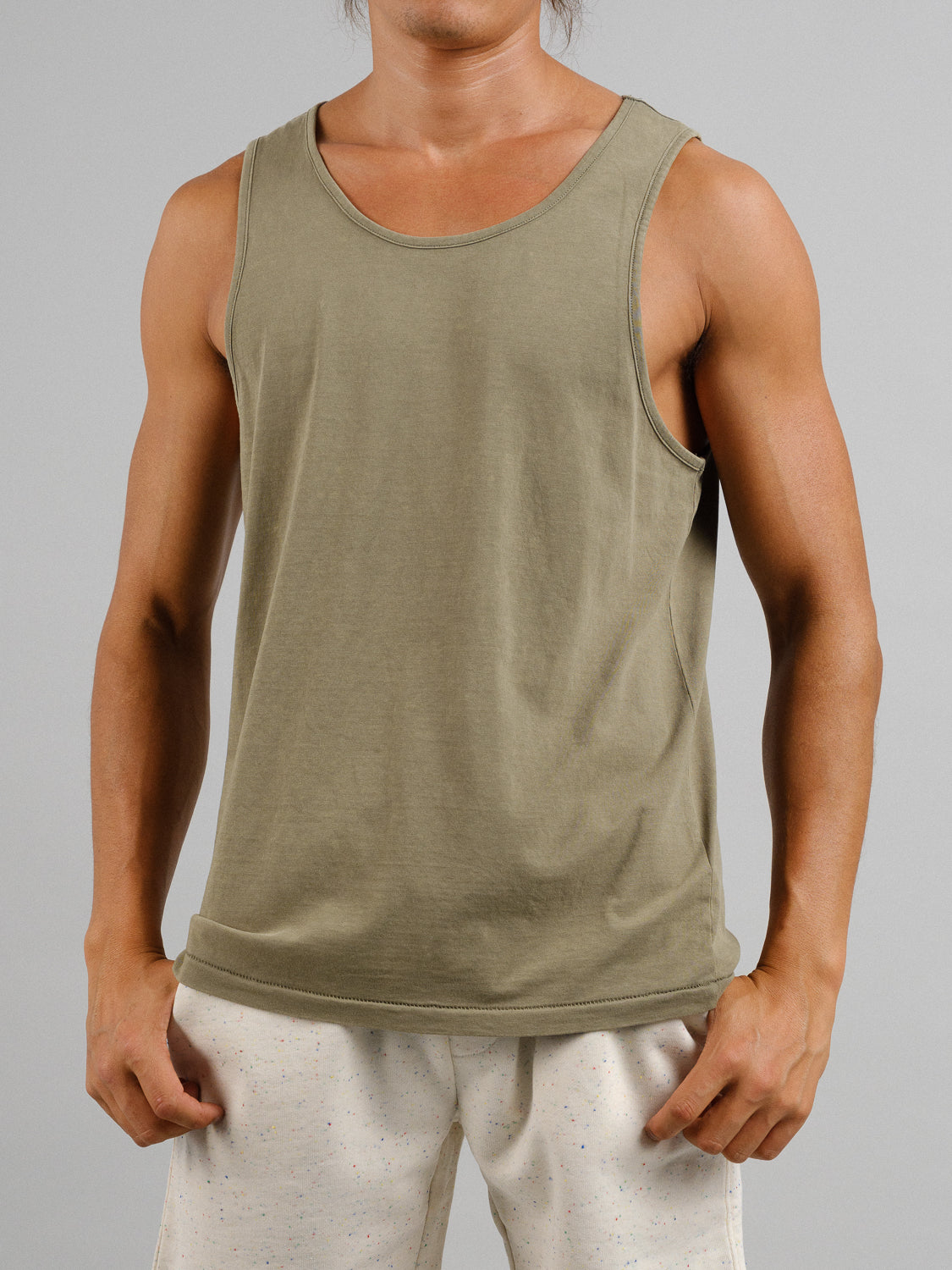 A person in a vintage-style ID Menswear Settler Snow Vintage Wash Tank Top, sleeveless and olive green, paired with white speckled shorts, poses against a plain gray background. Their toned arms rest casually near the shorts, enhancing the timeless look.